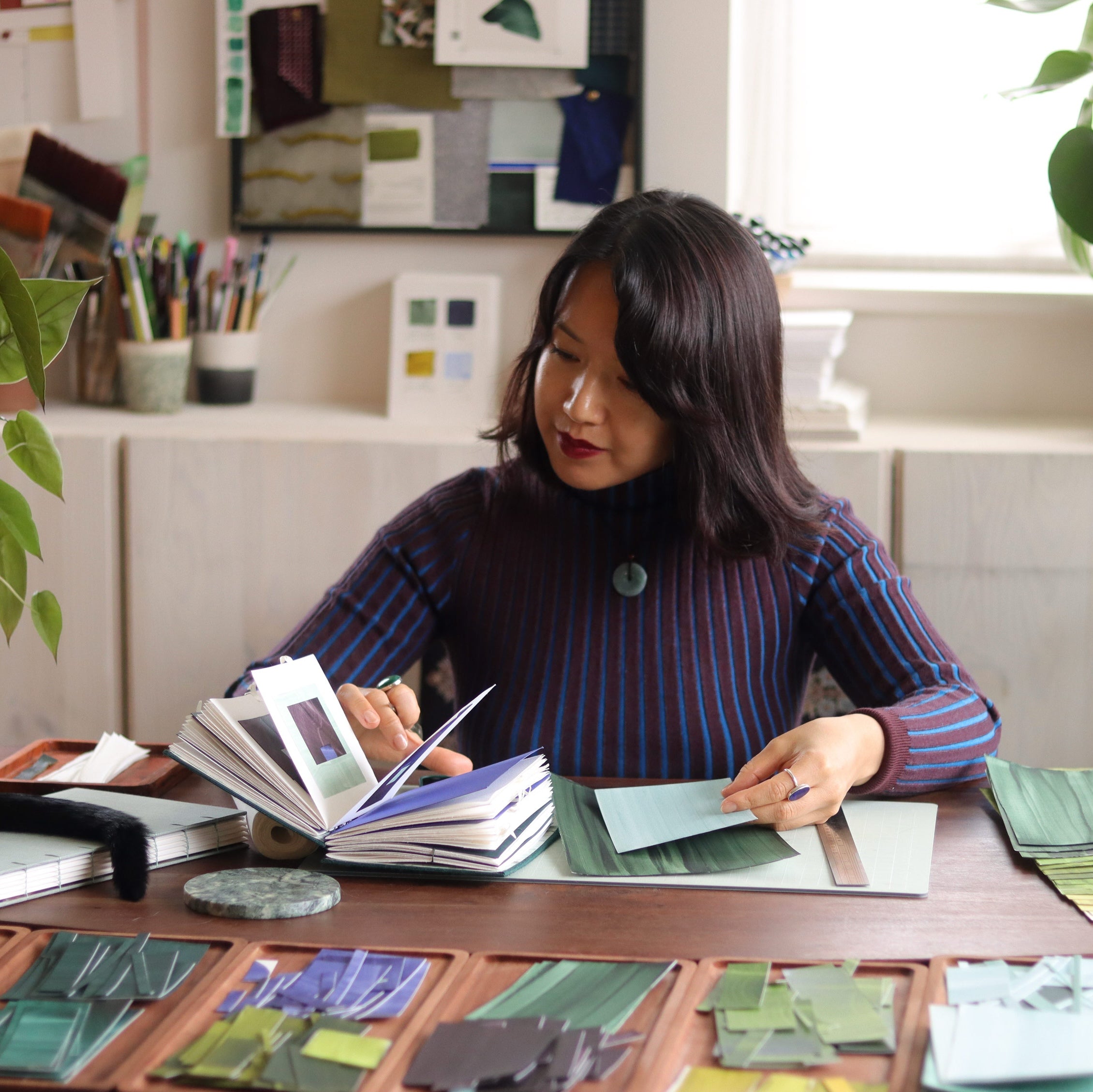 London based abstract artist Aimee Johanan sitting at her desk in the studio, with a sketchbook of artwork. Surrounded by painted papers in blues and greens, plants and art materials, while she works on some new abstract artwork.