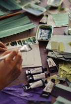 Creative messy studio workspace of the artist Aimee Johanan, with paint swatches, tubes of watercolour paint and the artist painting on paper.