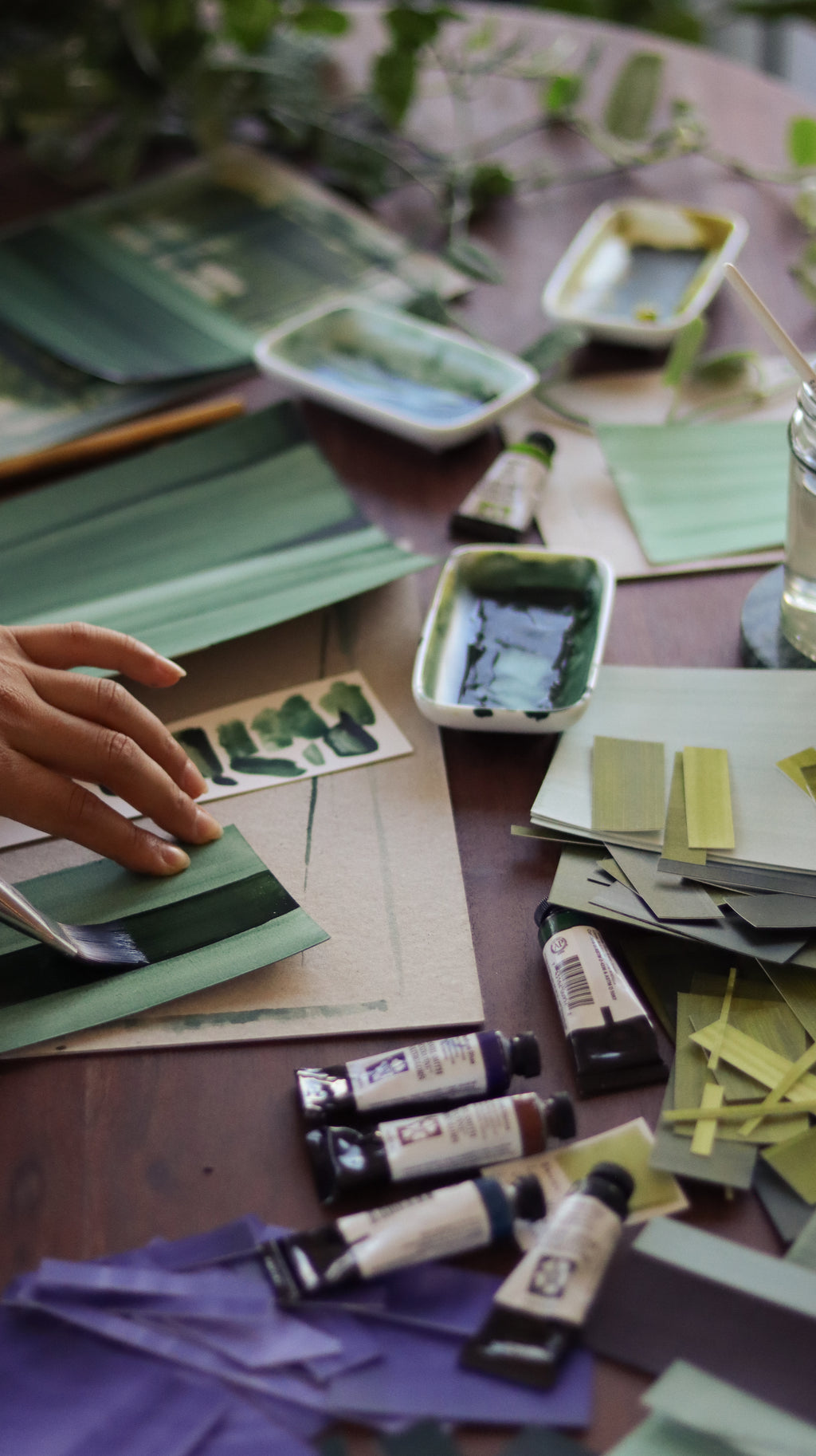Creative messy studio workspace of the artist Aimee Johanan, with paint swatches, tubes of watercolour paint and the artist painting on paper.