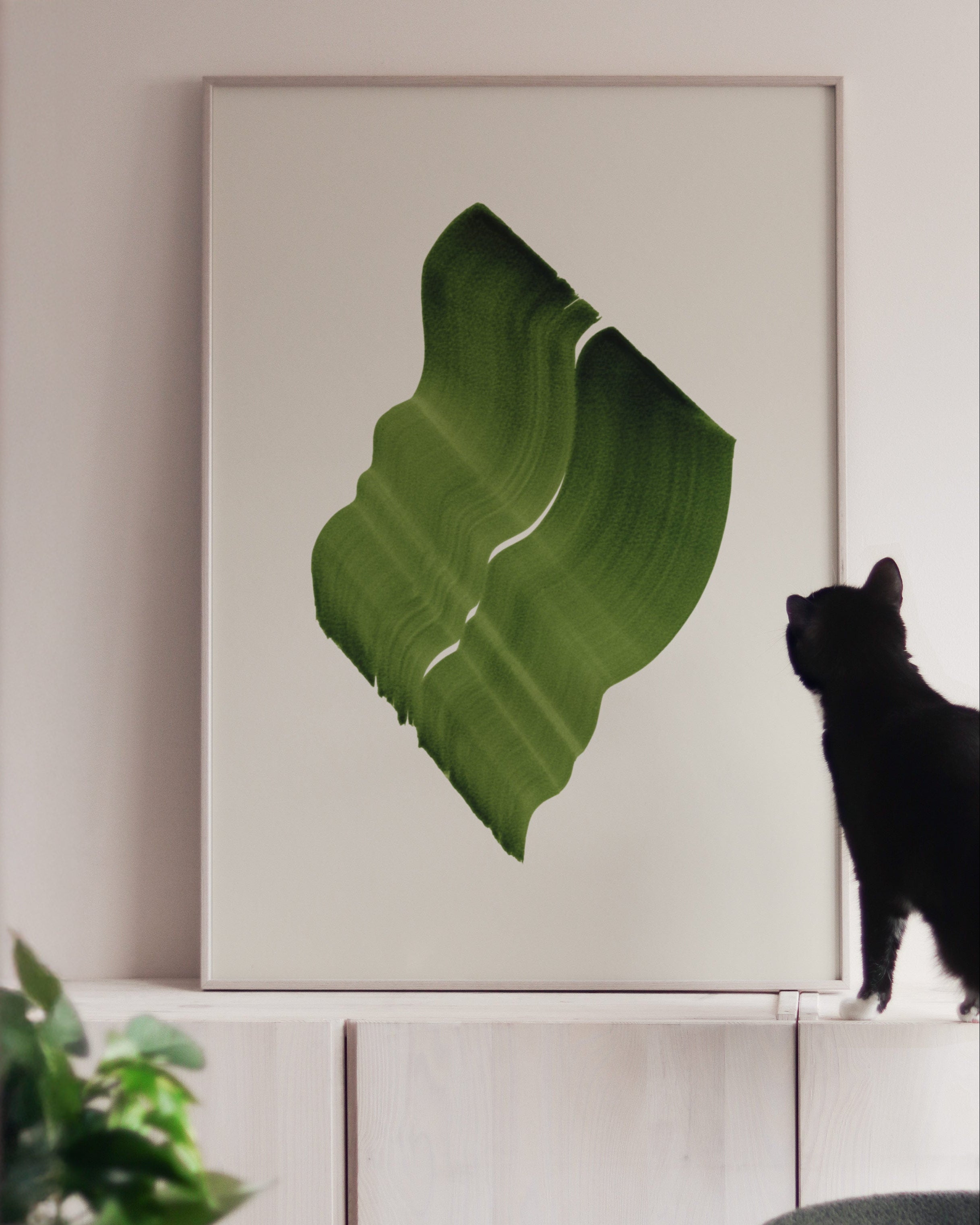 Large green art print in a white oak frame, on white cabinets with a black cat looking at it