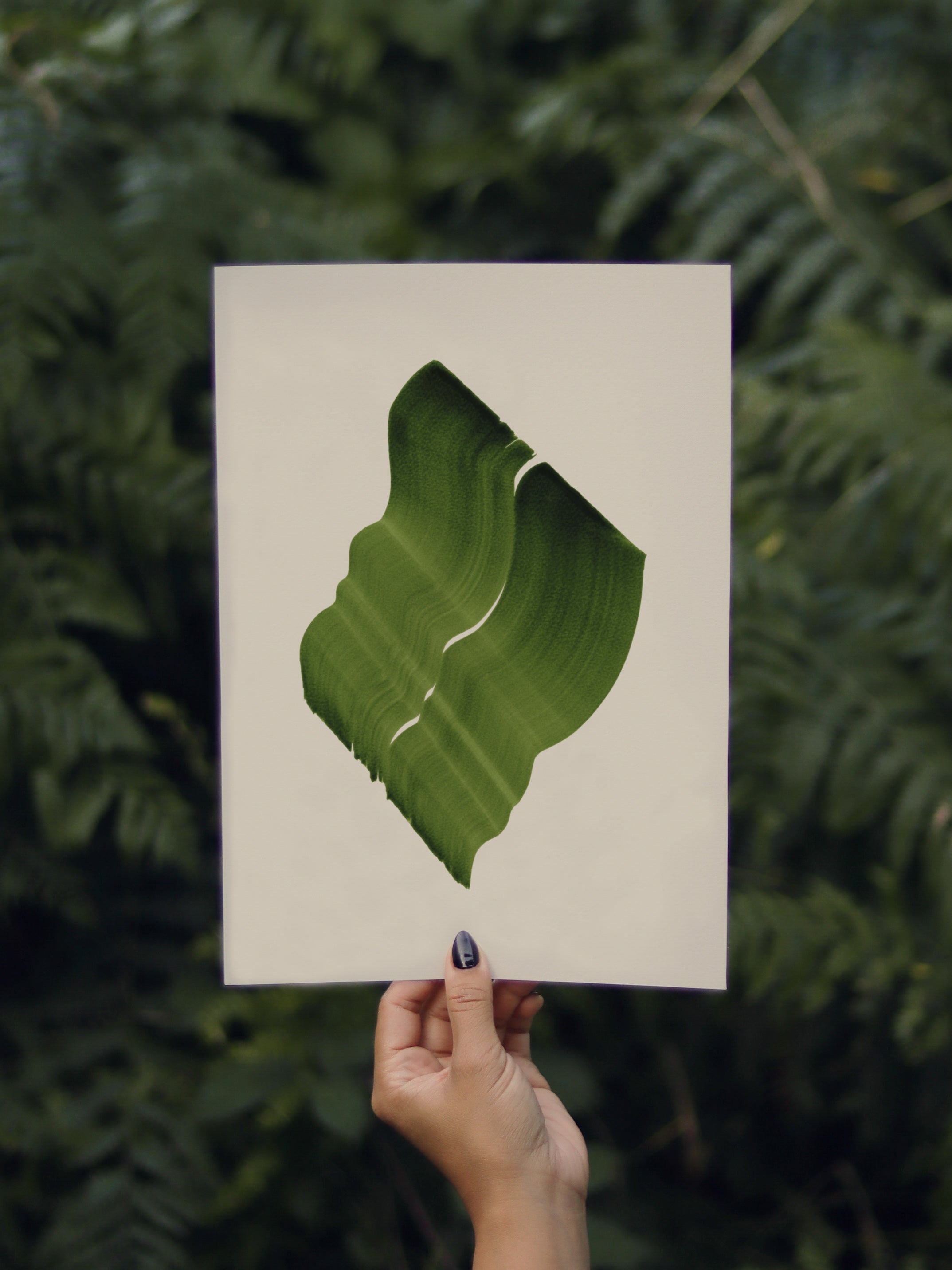 Hand holding a green abstract leaf-shaped print, against a green leafy background in the forest