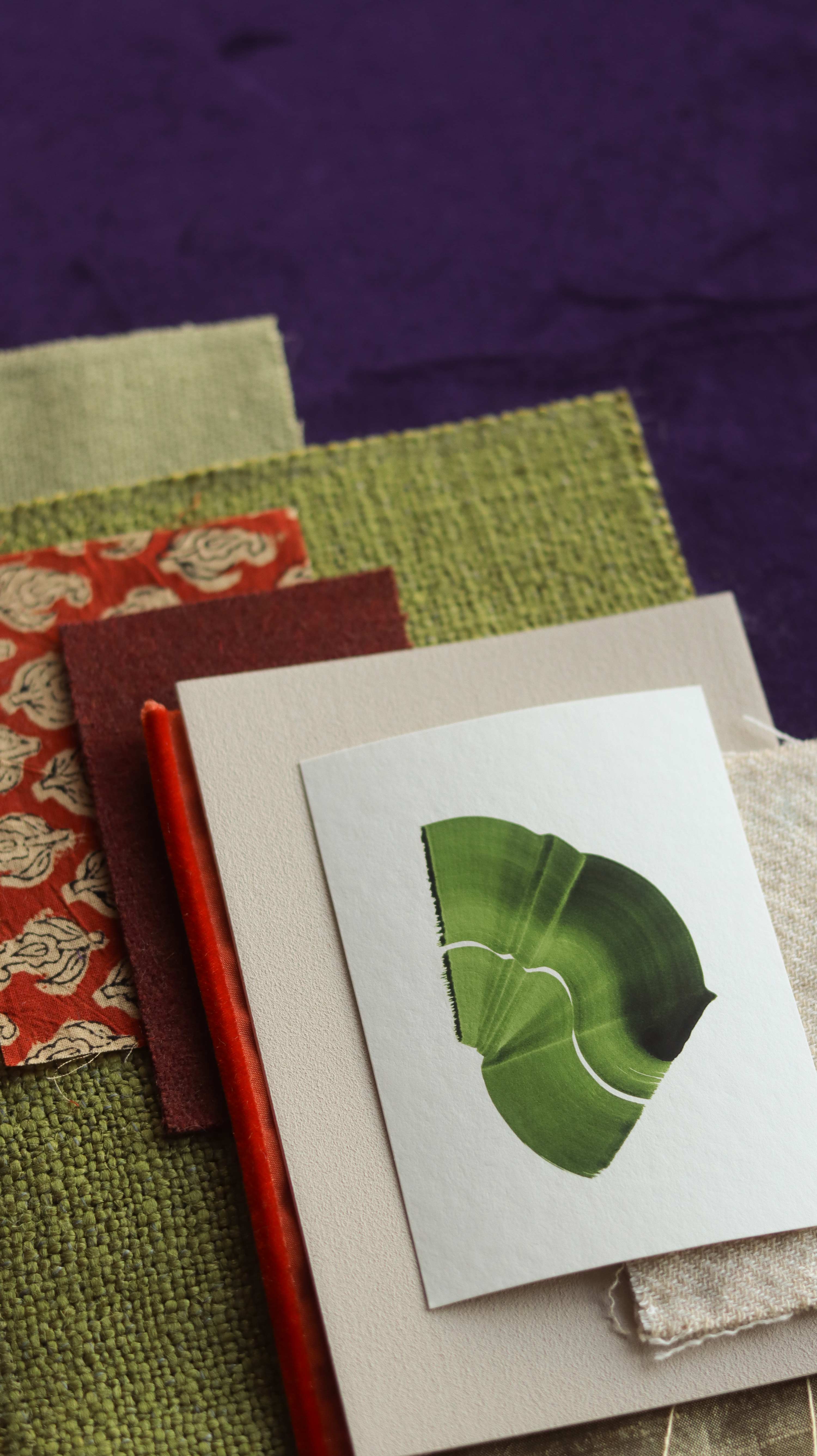 Green abstract art print sample with a green leaf-like design on a textured paint swatch with fabric swatches. Inspiration for green orange and purple themed interior decor.