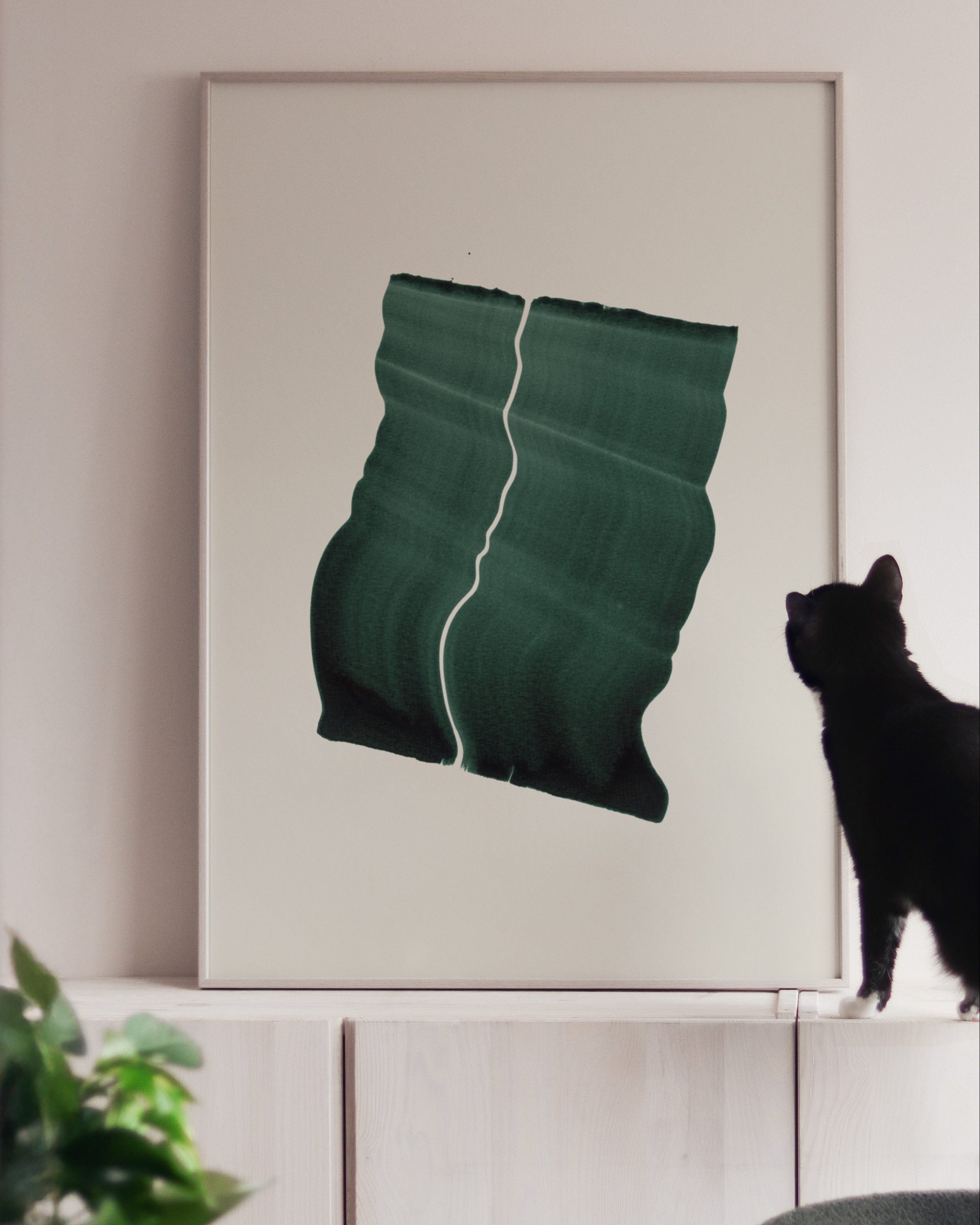 Large green art print in a white oak frame, on white cabinets with a black cat looking at it