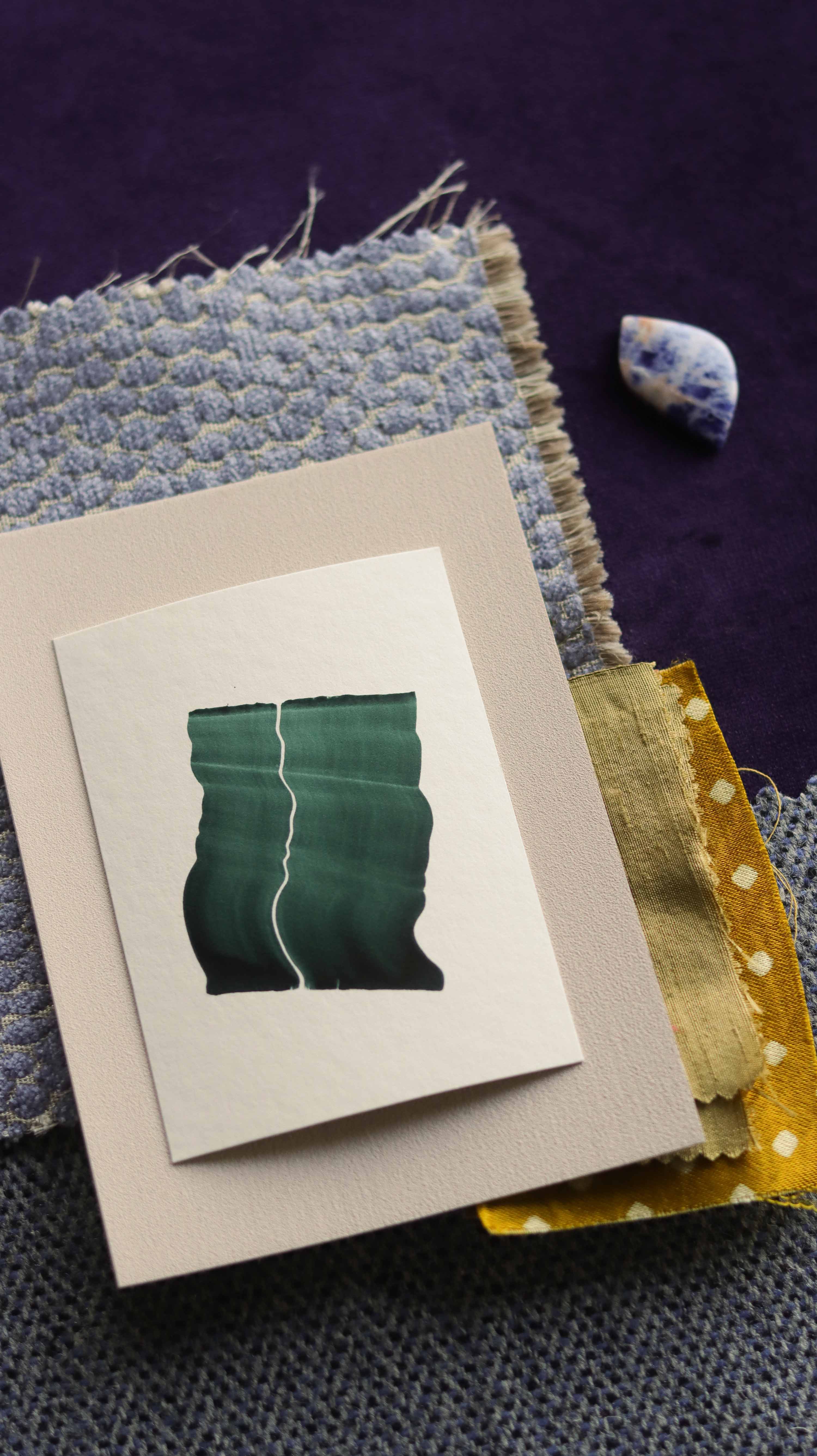 dark green abstract art print sample with a green leaf-like design on a textured paint swatch with fabric swatches. Inspiration for dusty blue, ink purple and earth tone themed interior design.