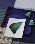 Dark green abstract art print sample with a green leaf-like design laying on top of a textured paint swatch with fabric swatches and green malachite stones. Inspiration for green and blue themed interior decor.