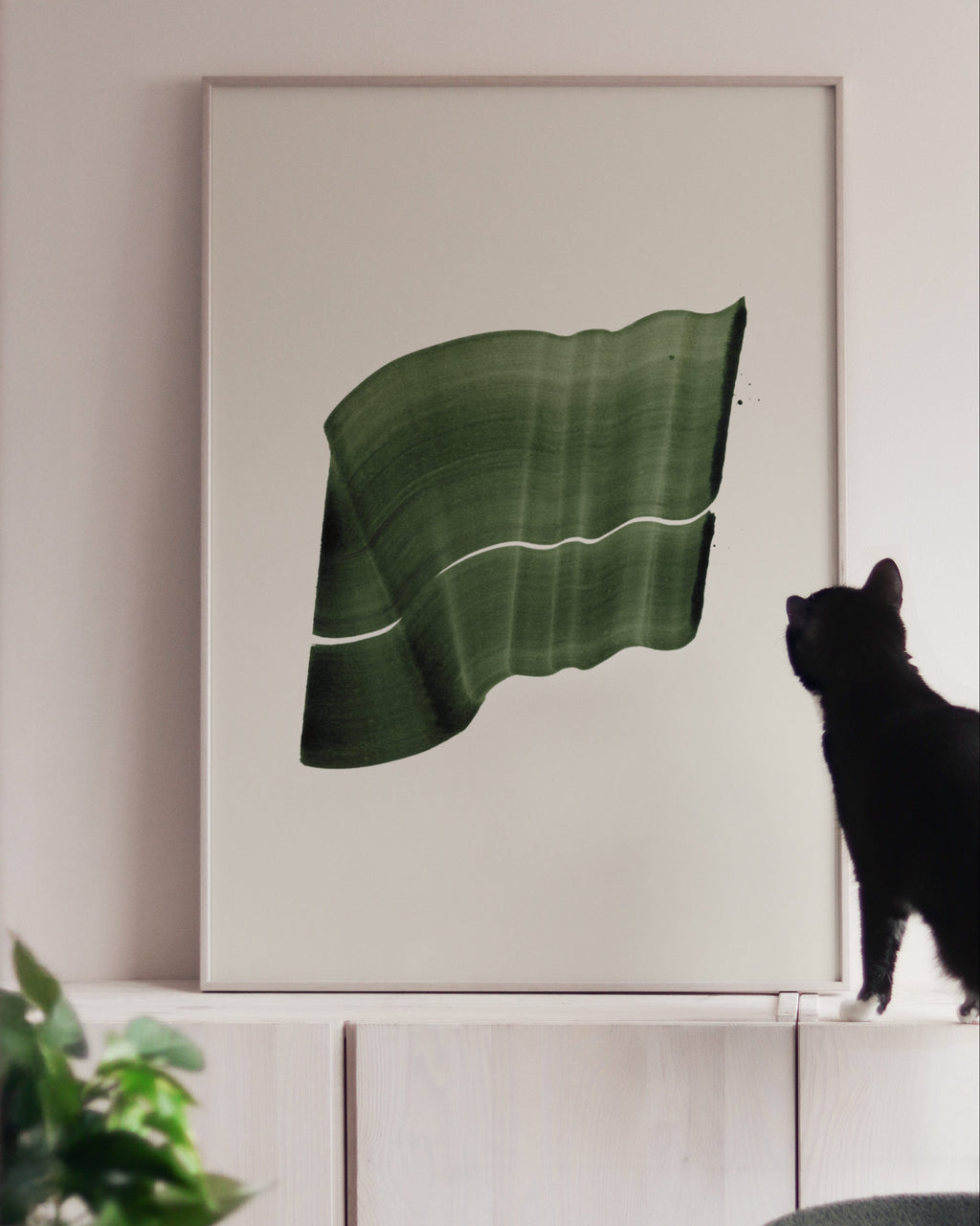 Large green art print in a white oak frame, on white cabinets with a black cat looking at it
