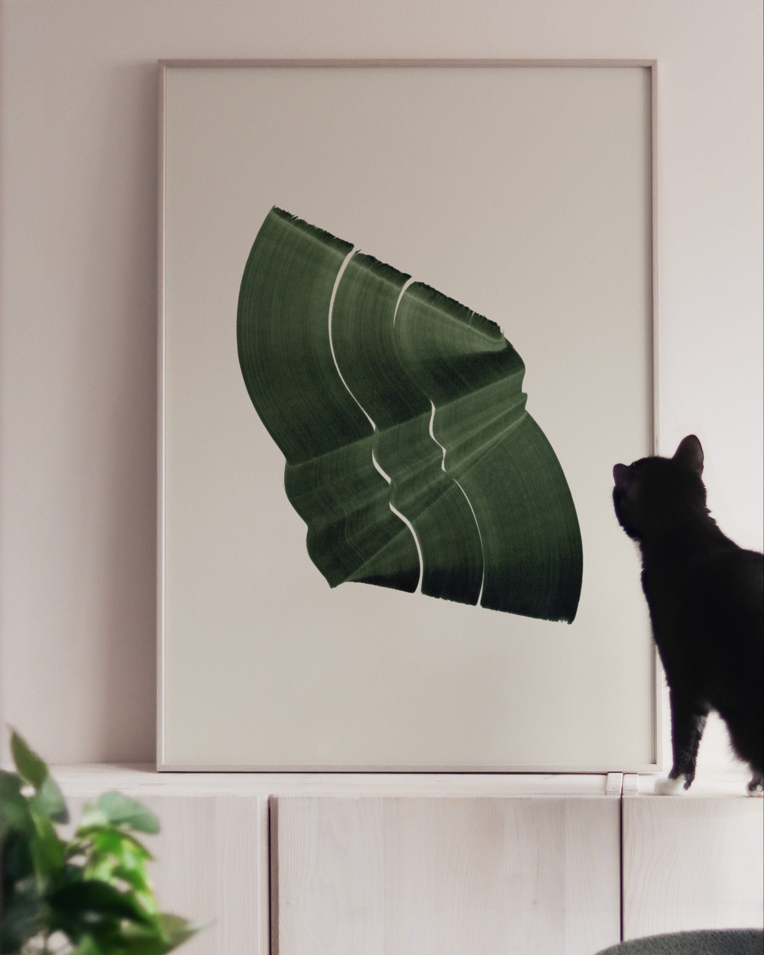 Large green art print in a white oak frame, on white cabinets with a black cat looking at it