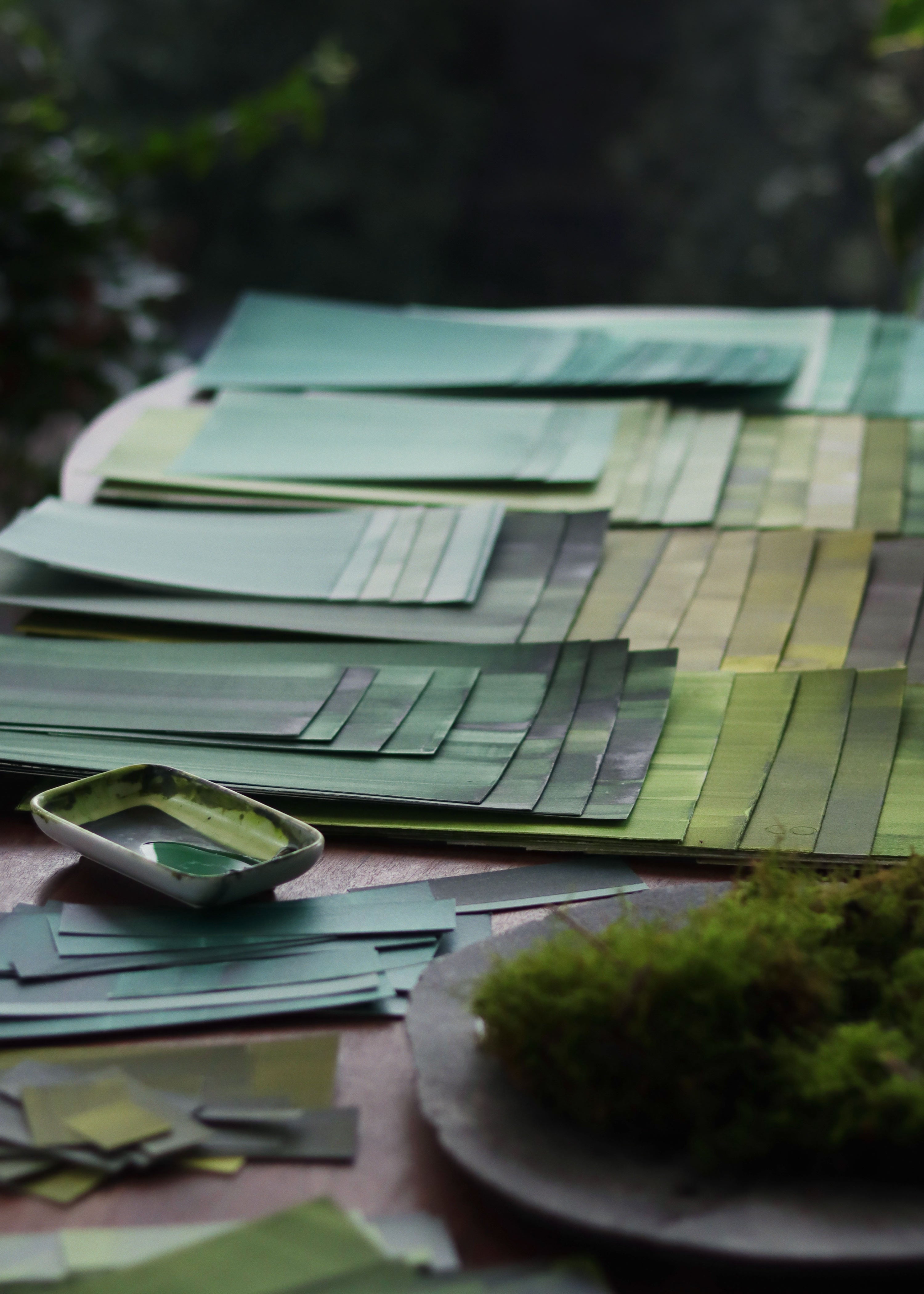 Green and blue painted papers laid out on a desk with paint and moss, in London based abstract artist Aimee Johanan's creative studio.
