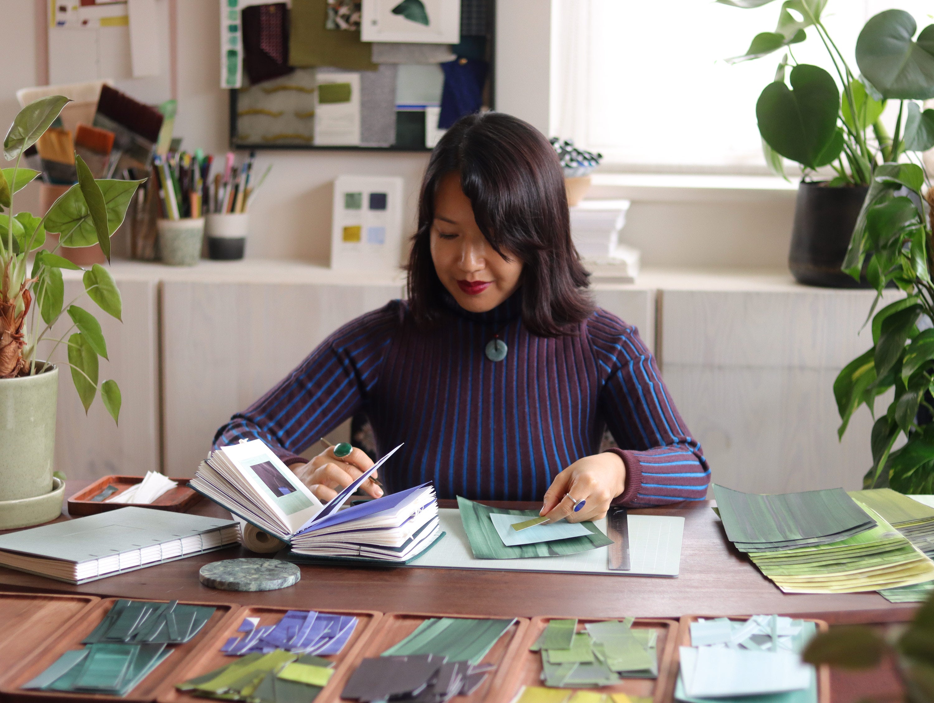 London based abstract artist Aimee Johanan working at her desk in her creative studio, surrounded by materials, sketchbooks and plants