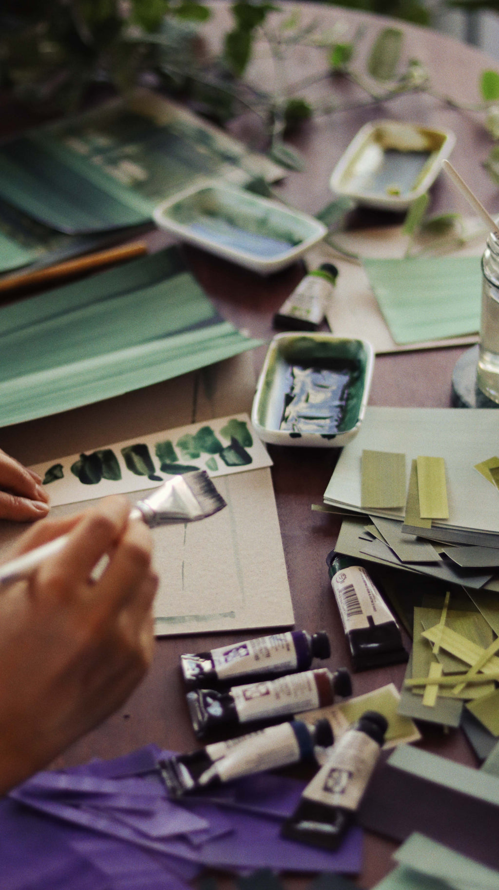 Messy table with works in progress in abstract artist Aimee Johanan's studio in London, covered in paint swatches, tubes of watercolour and painted papers.