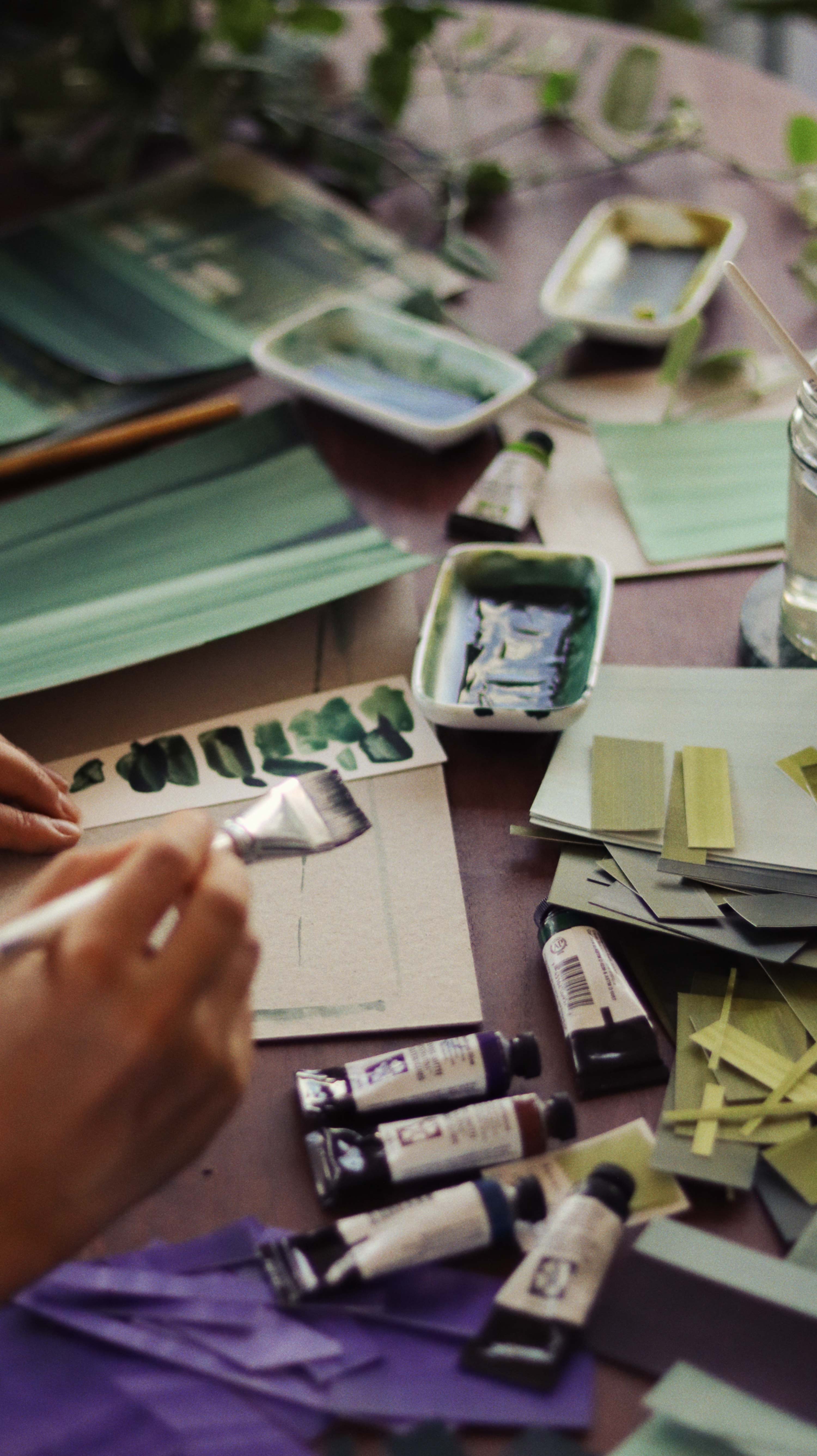 Messy table with works in progress in abstract artist Aimee Johanan's studio in London, covered in paint swatches, tubes of watercolour and painted papers.