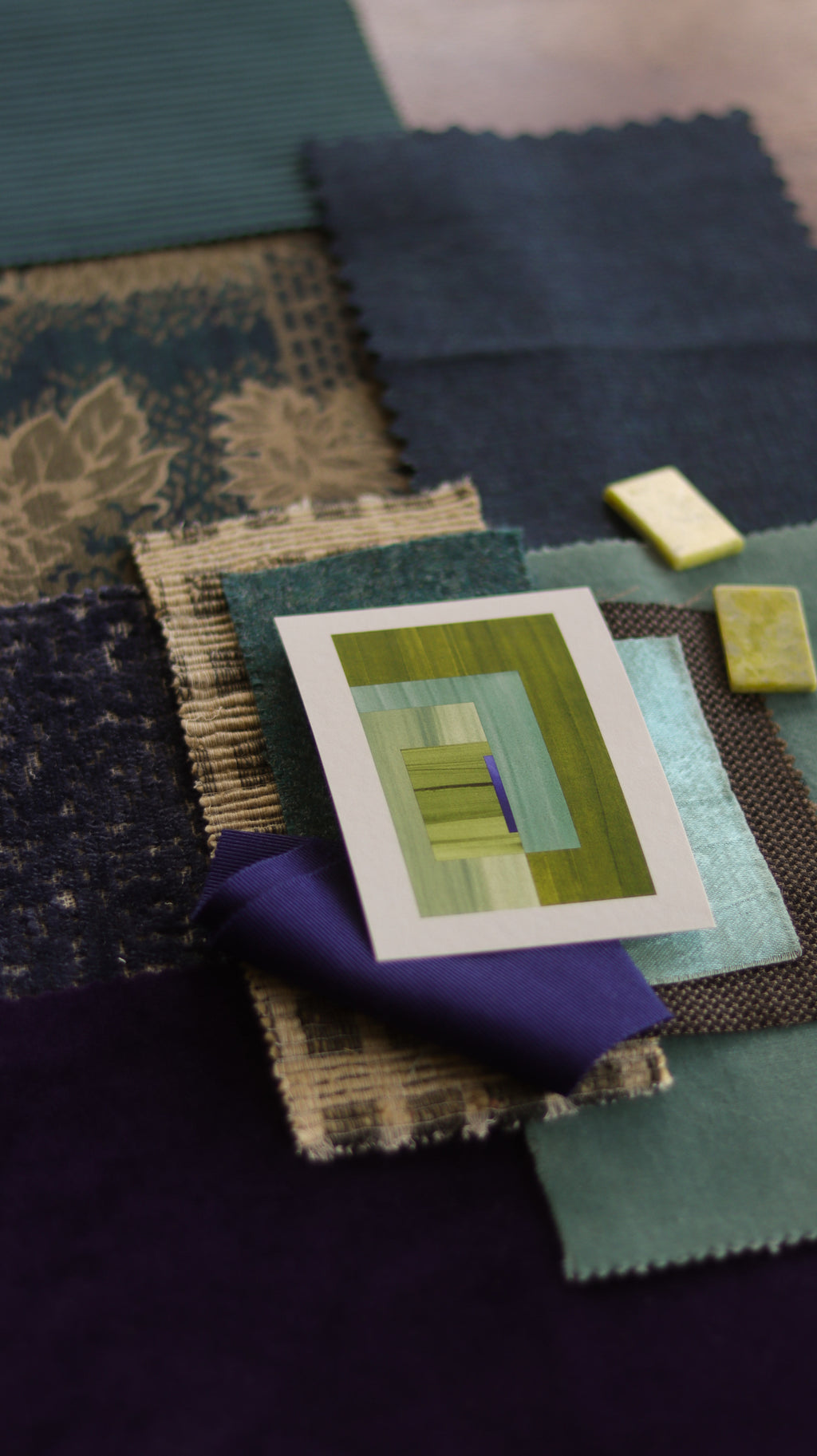 Small abstract art print sample with moss green and blue colours, laying on top of fabric swatches for a blue and teal interior colour scheme.