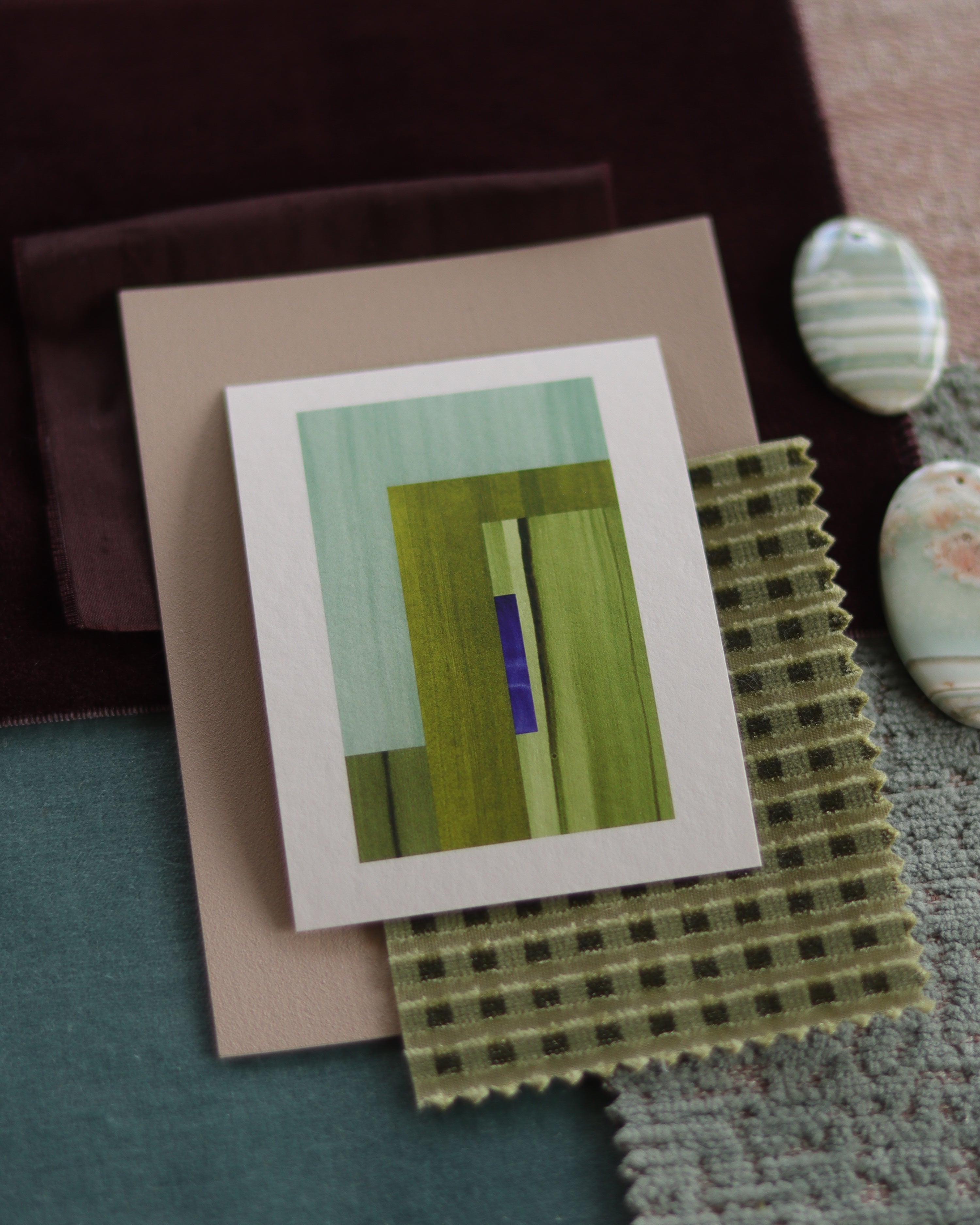 Small abstract art print sample with moss green, turquoise and blue colours, laying on top of fabric swatches for interiors colour inspiration.