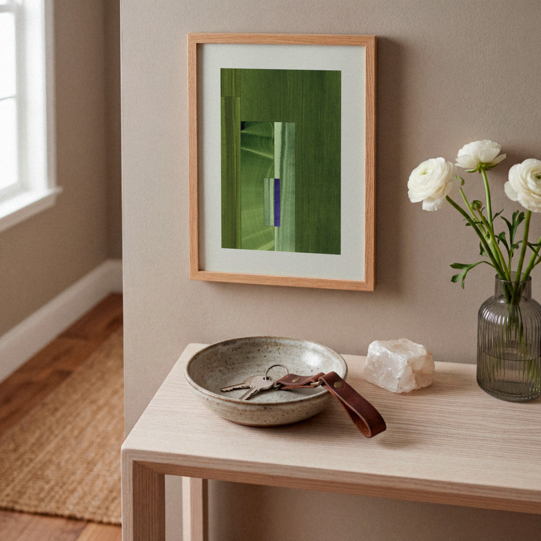 Framed green abstract artwork on a white oak console table, in a modern hallway, with a case of white flowers