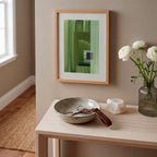 Framed green abstract artwork on a white oak console table, in a modern hallway, with a case of white flowers