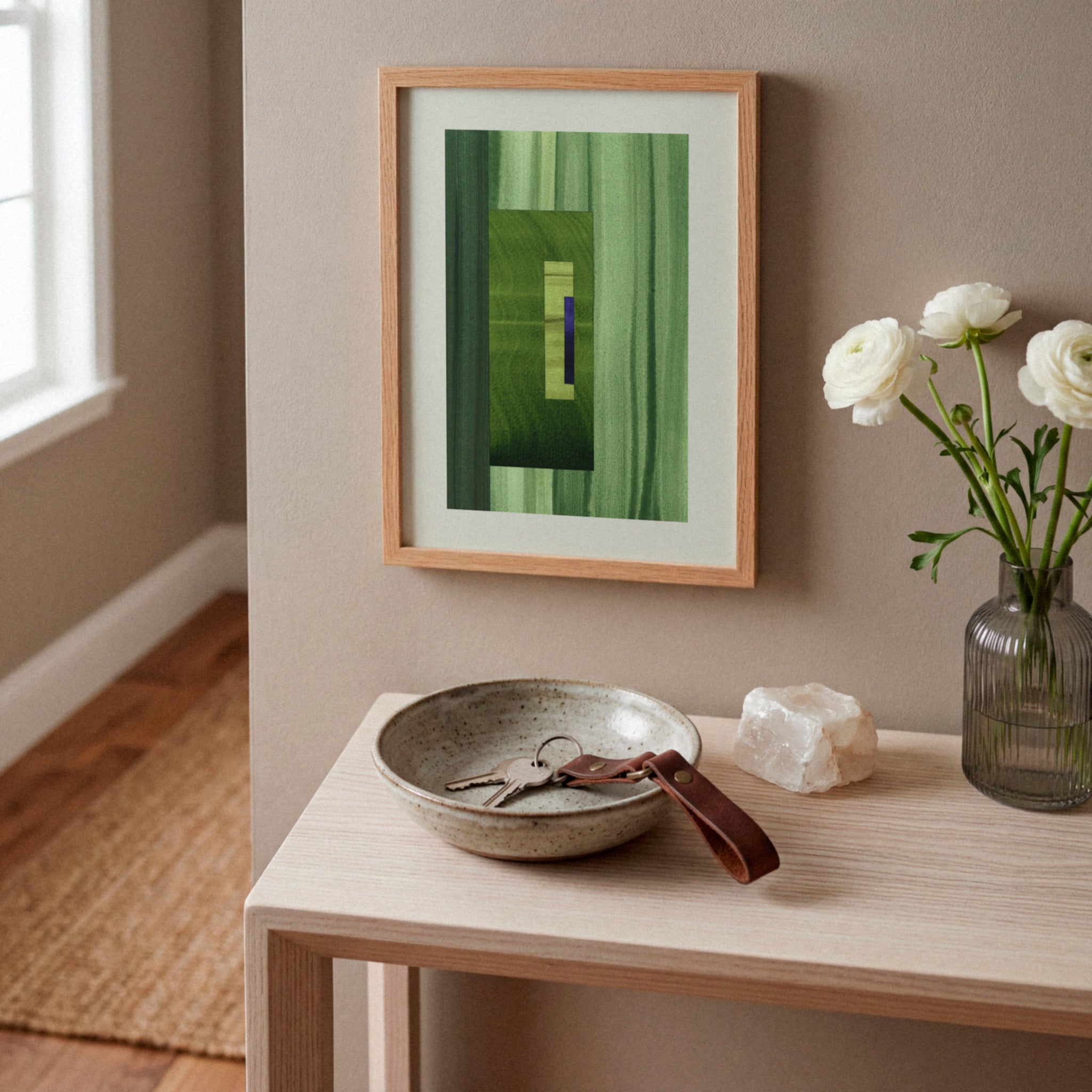 Framed green abstract artwork on a white oak console table, in a modern hallway, with a case of white flowers