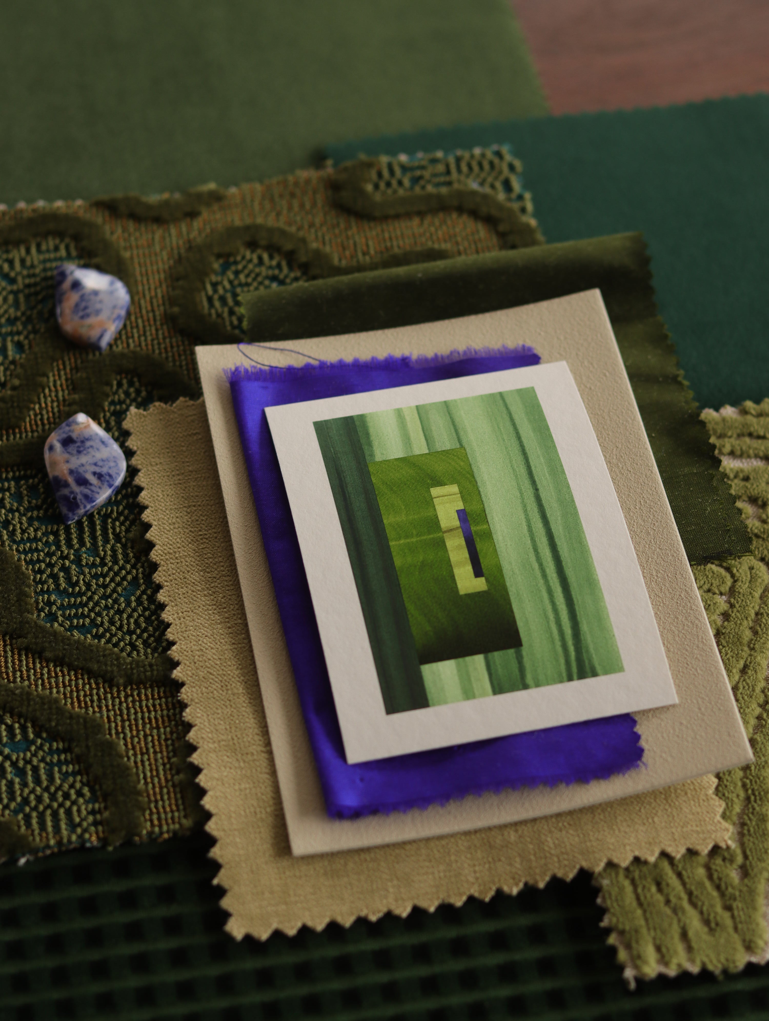 Small abstract art print sample with moss green, grass green and blue colours, laying on top of fabric swatches for green interiors colour inspiration.