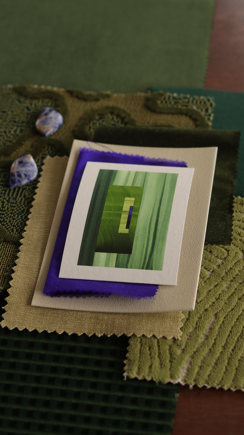 Small abstract art print sample with moss green, grass green and blue colours, laying on top of fabric swatches for green interiors colour inspiration.