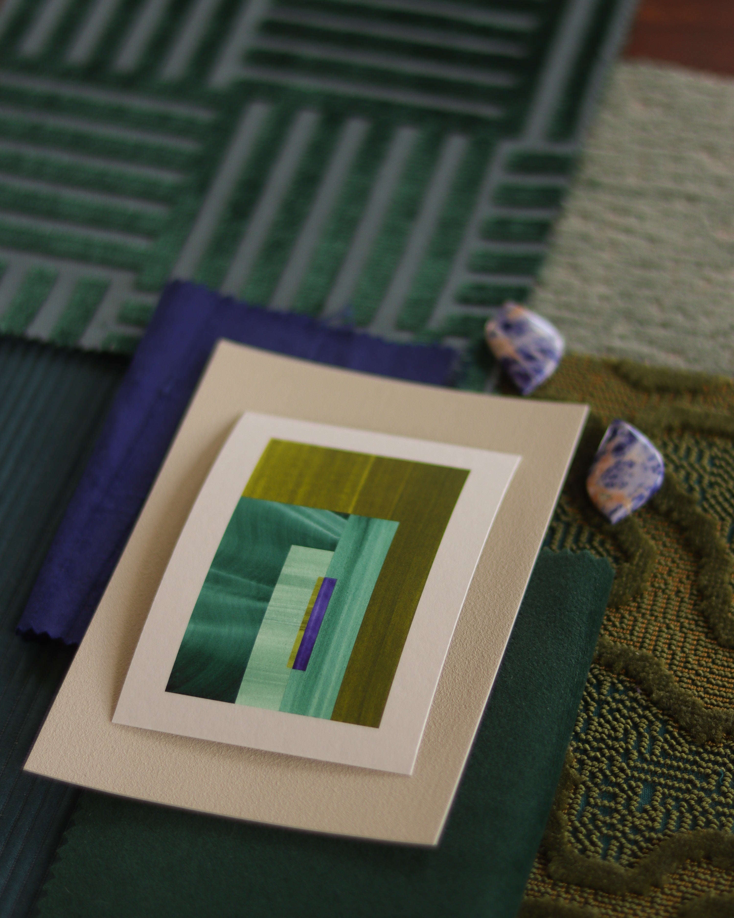 Small geometric abstract art print sample with forest green, moss green  and blue colours, laying on top of fabric swatches for colourful interiors inspiration.