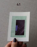 Hand holding sample of a turquoise, eggplant/aubergine purple and royal blue minimalist abstract art print with a geometric design over a grey linen background