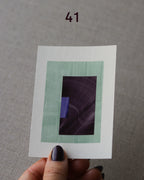 Hand holding sample of a turquoise, eggplant/aubergine purple and royal blue minimalist abstract art print with a geometric design over a grey linen background