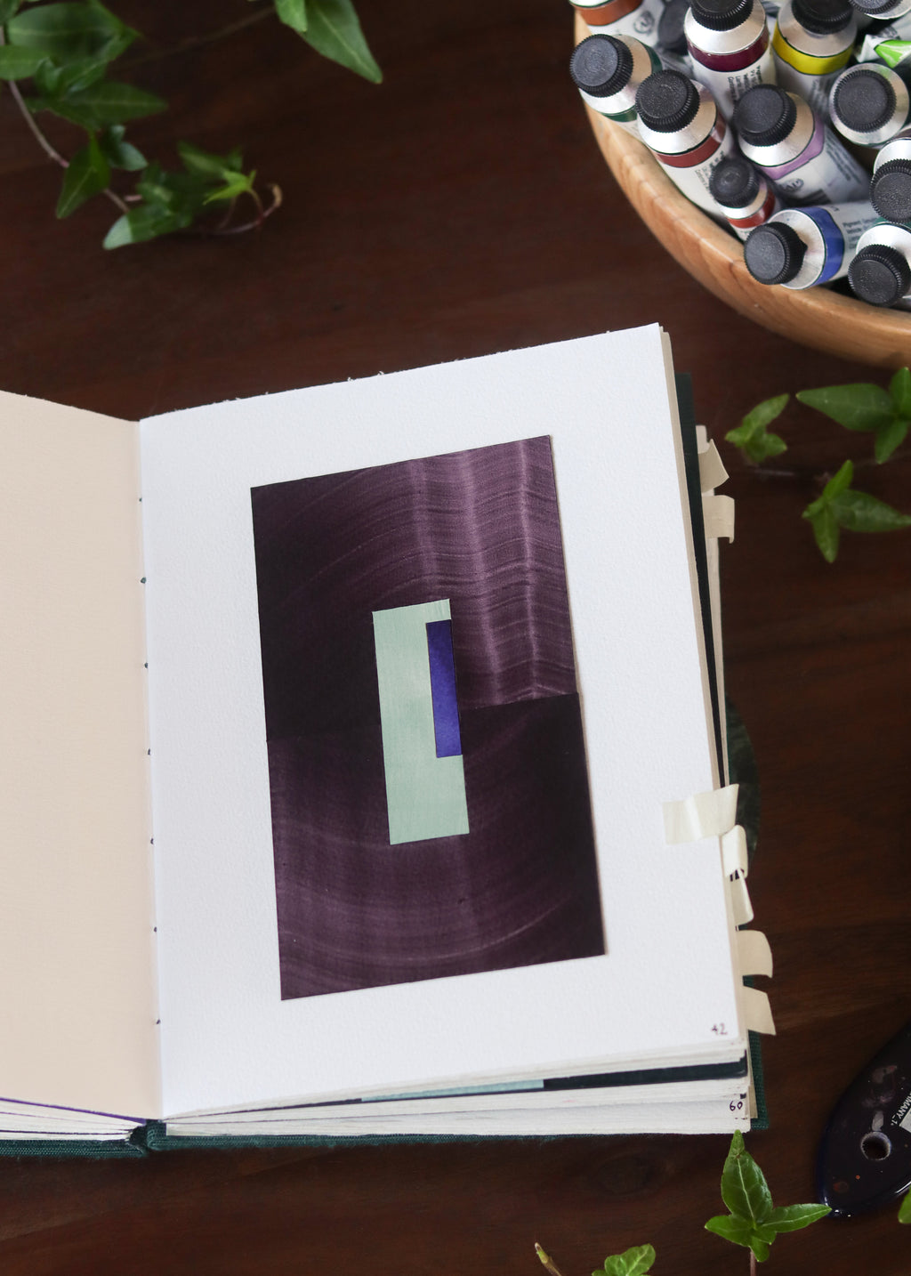Open sketchbook with a purple geometric abstract art piece on a dark wooden table with tubes of watercolour paint and plants.