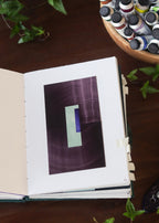 Open sketchbook with a purple geometric abstract art piece on a dark wooden table with tubes of watercolour paint and plants.