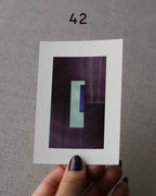 Hand holding sample of a turquoise, eggplant/aubergine purple and royal blue minimalist abstract art print with a geometric design over a grey linen background