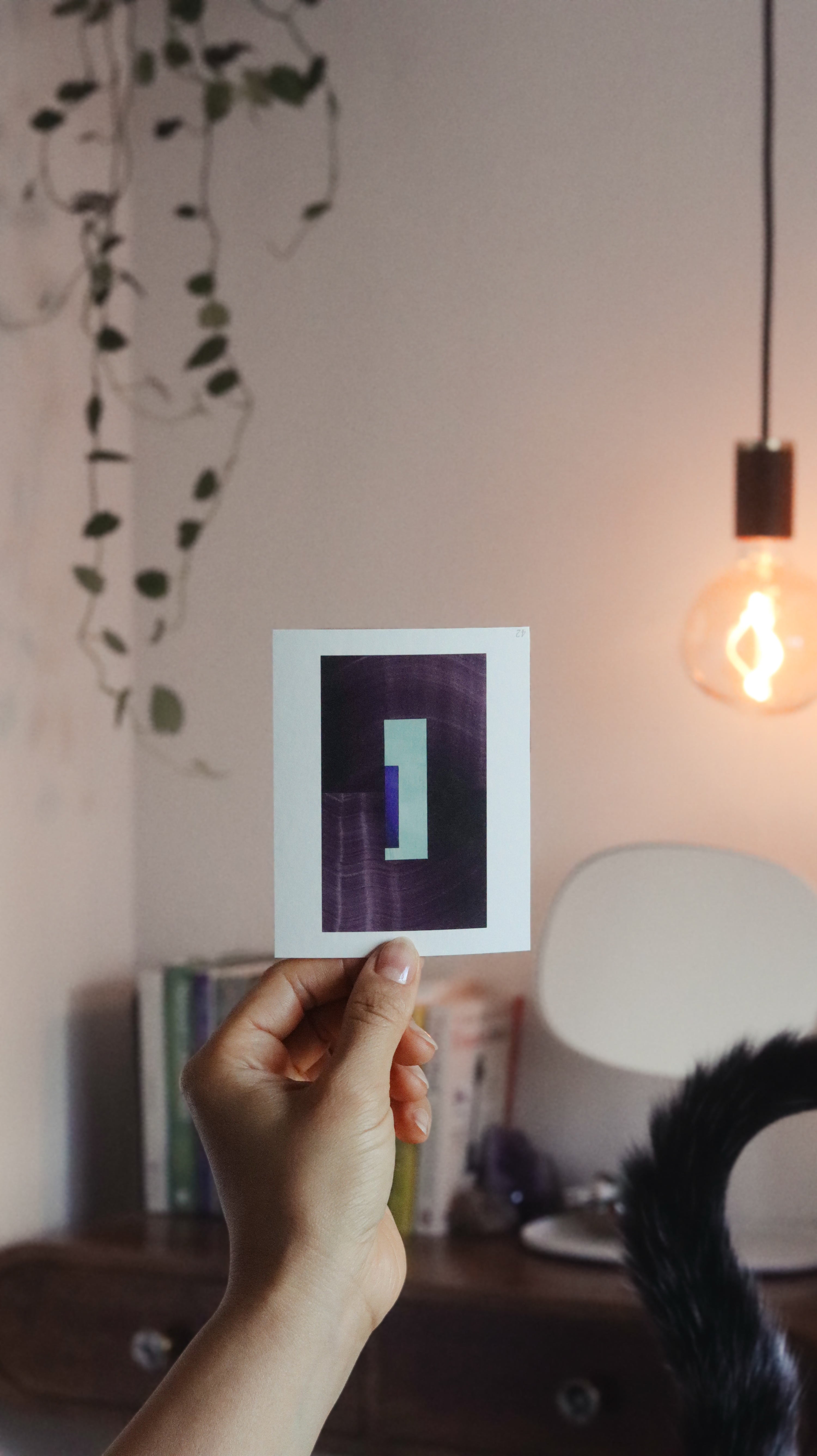 Hand holding a minimalist abstract art print sample with aubergine/eggplant purple and royal blue colours, in a peaceful, modern bedroom over the bedside table