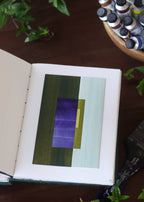 Open sketchbook with a geometric abstract art piece on a dark wooden table with tubes of watercolour paint and plants.
