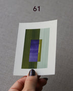 Hand holding sample of a green and blue minimalist abstract art print with a geometric design over a grey linen background