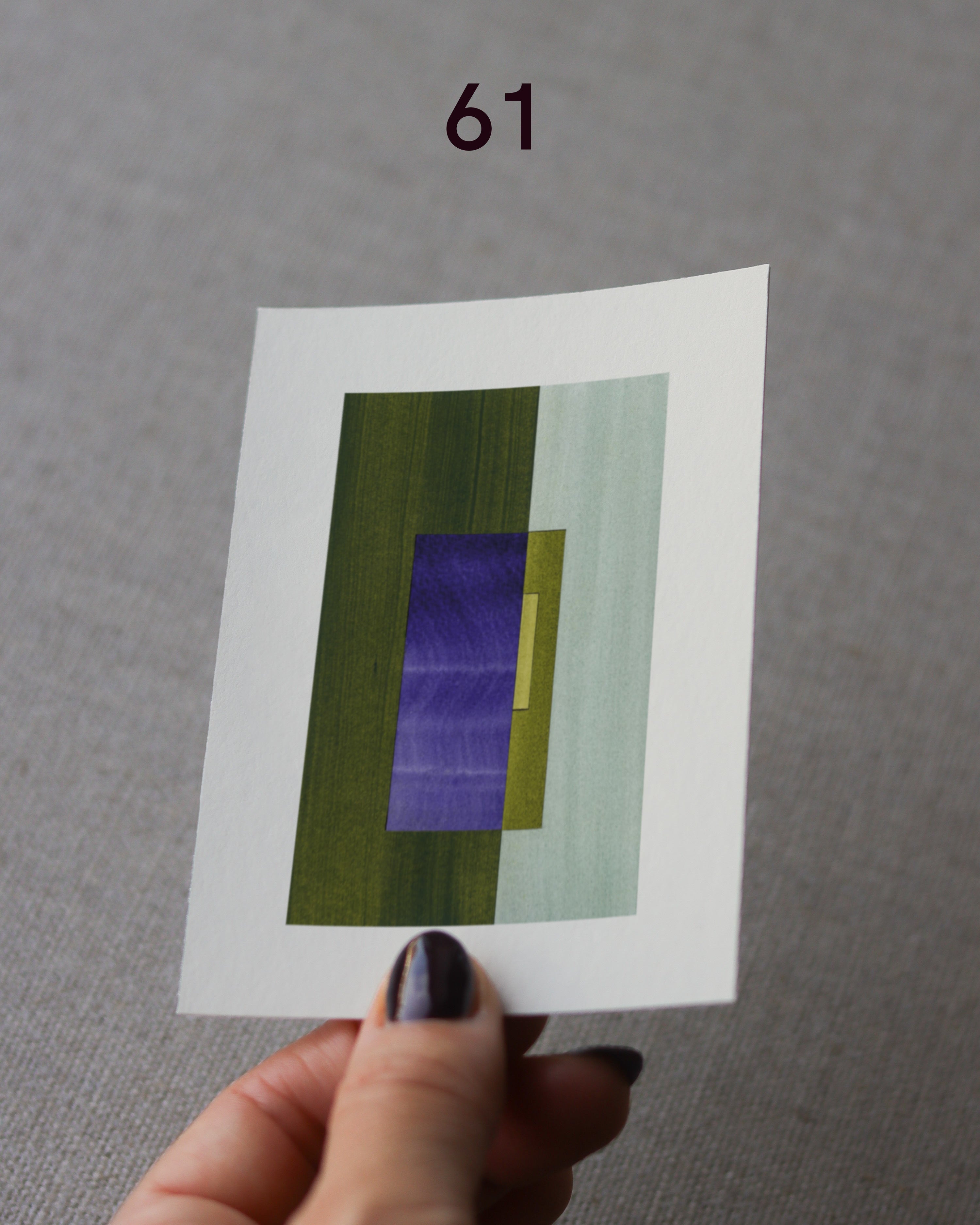 Hand holding sample of a green and blue minimalist abstract art print with a geometric design over a grey linen background