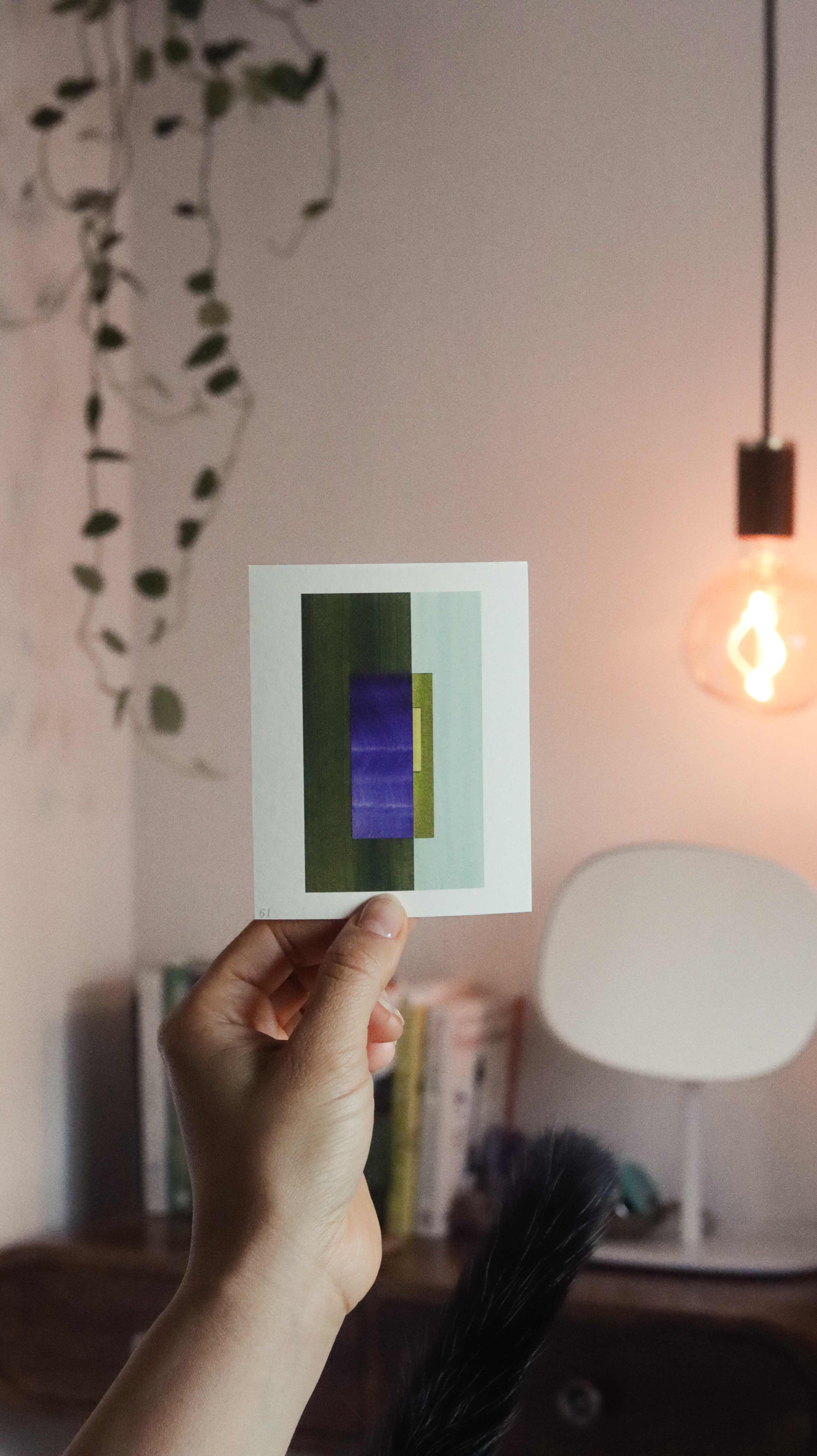 Hand holding up a blue and green geometric abstract art print sample in a peaceful, modern bedroom with plants and pendant lights