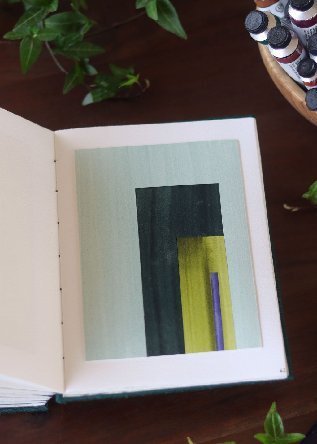 Open sketchbook with a geometric abstract art piece on a dark wooden table with tubes of watercolour paint and plants.