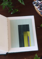 Open sketchbook with a geometric abstract art piece on a dark wooden table with tubes of watercolour paint and plants.