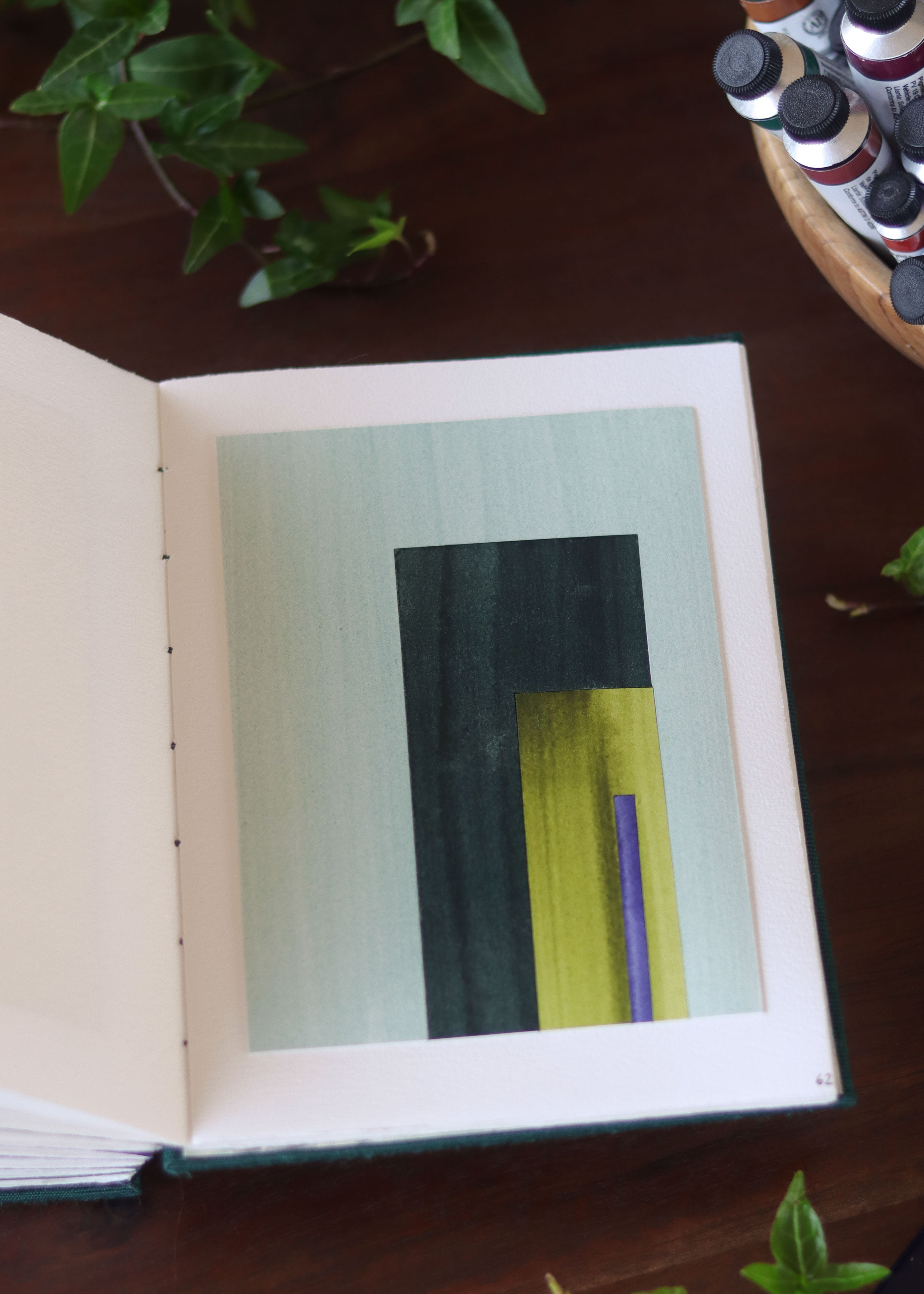 Open sketchbook with a geometric abstract art piece on a dark wooden table with tubes of watercolour paint and plants.