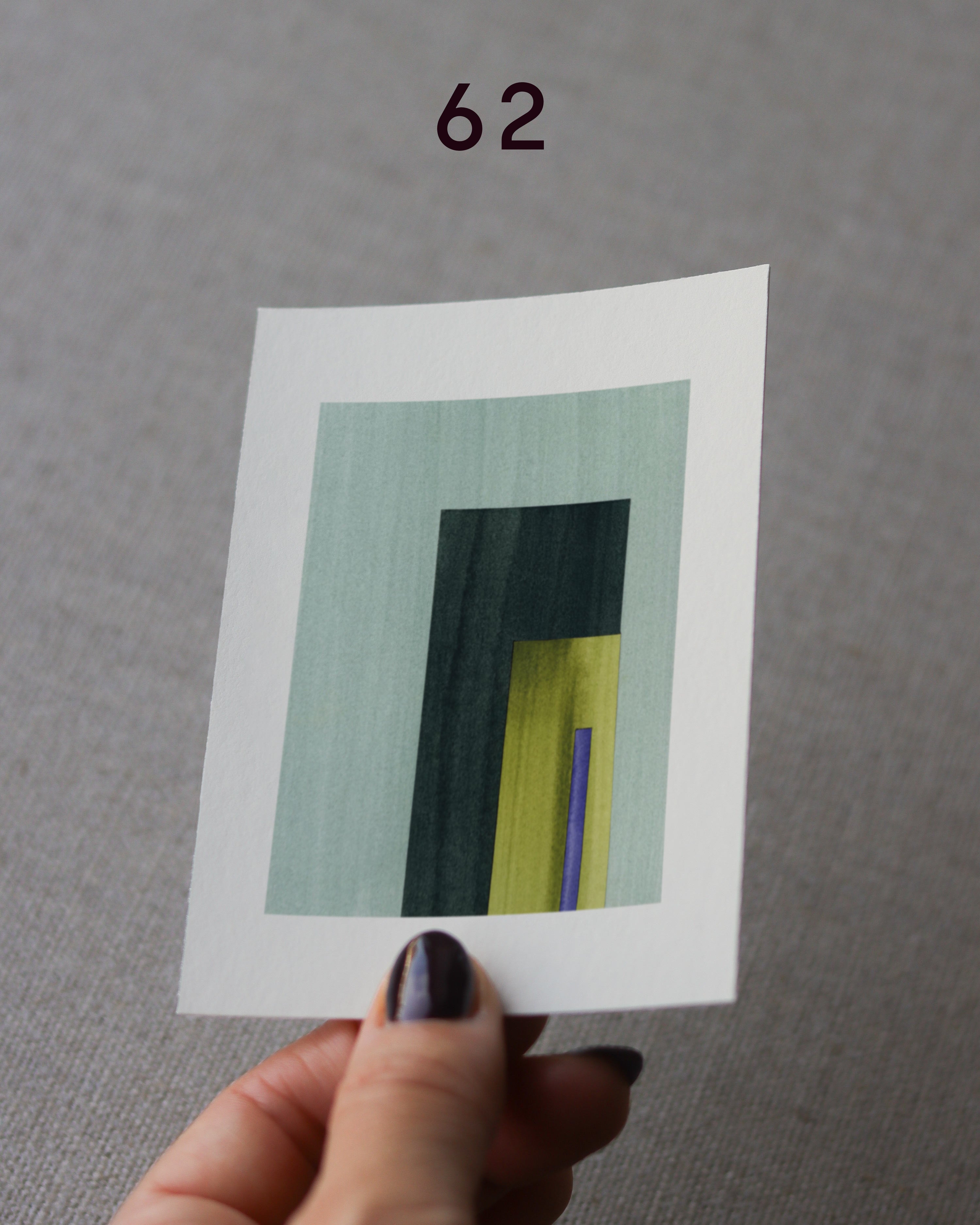 Hand holding sample of a turquoise,green, yellow and blue minimalist abstract art print with a geometric design over a grey linen background