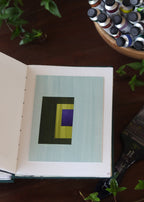 Open sketchbook with a geometric abstract art piece on a dark wooden table with tubes of watercolour paint and plants.