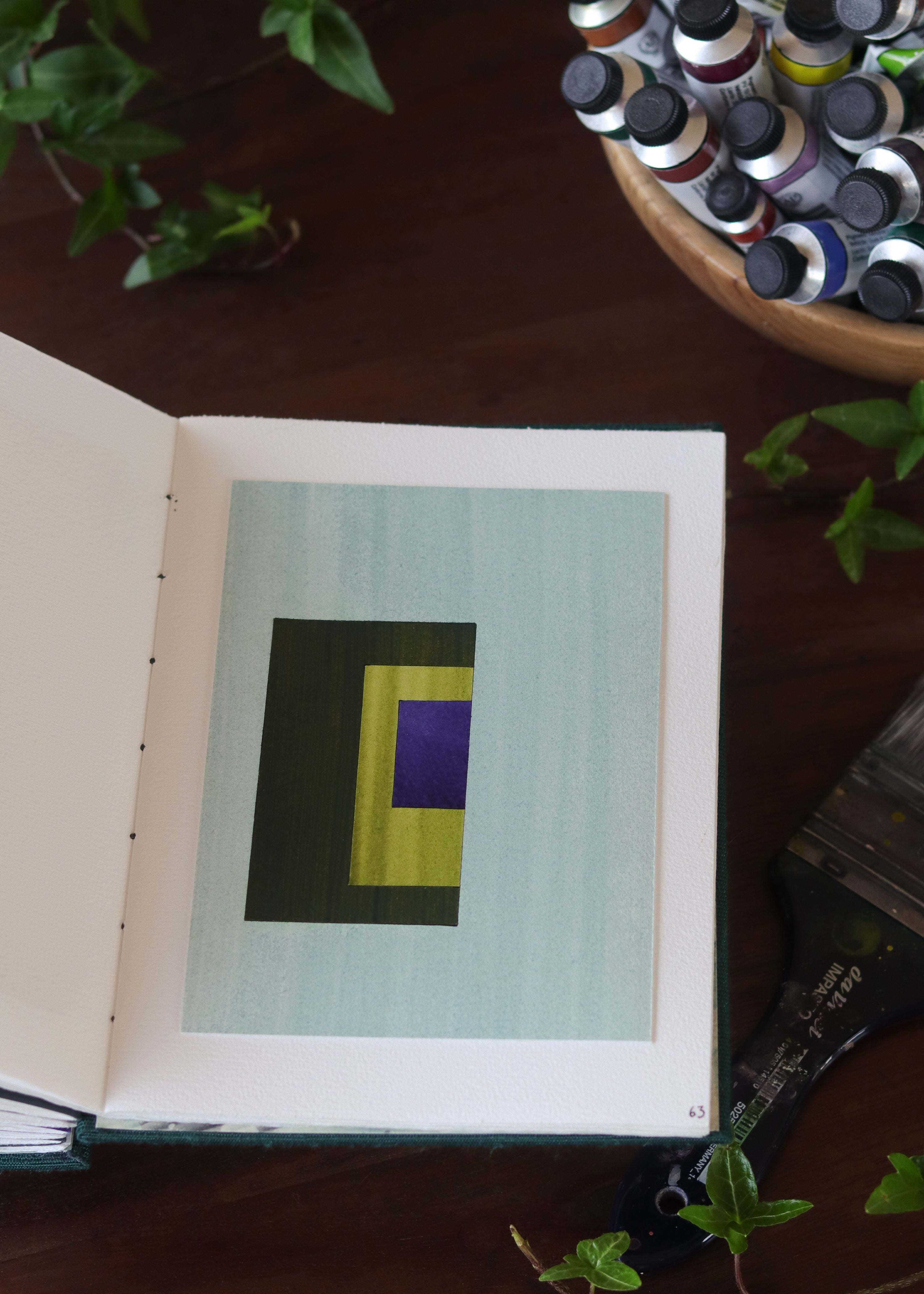 Open sketchbook with a geometric abstract art piece on a dark wooden table with tubes of watercolour paint and plants.