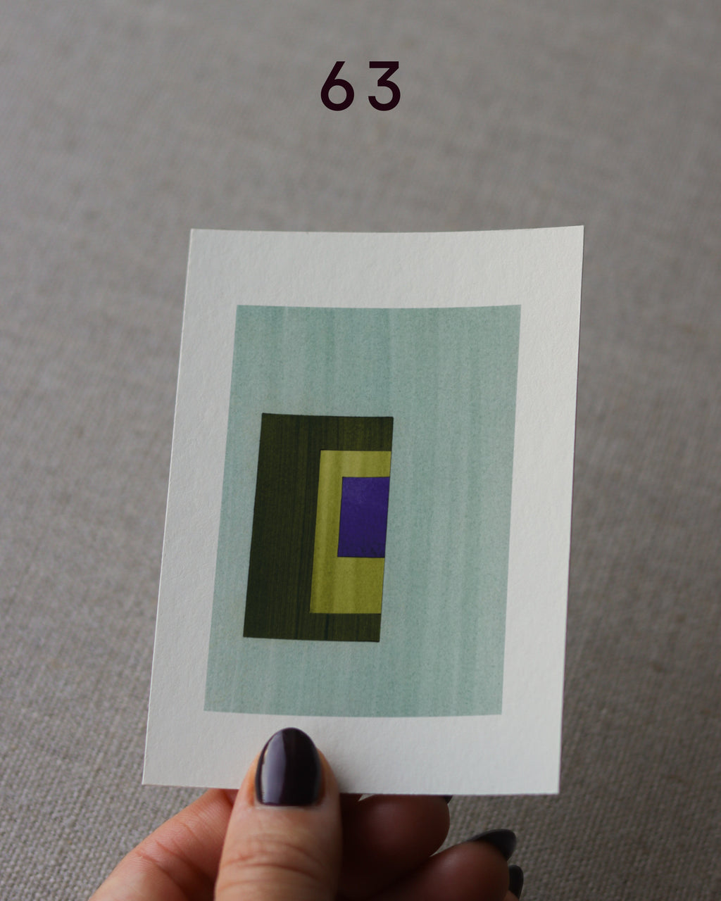 Hand holding sample of a turquoise, olive green, yellow and blue minimalist abstract art print with a geometric design over a grey linen background