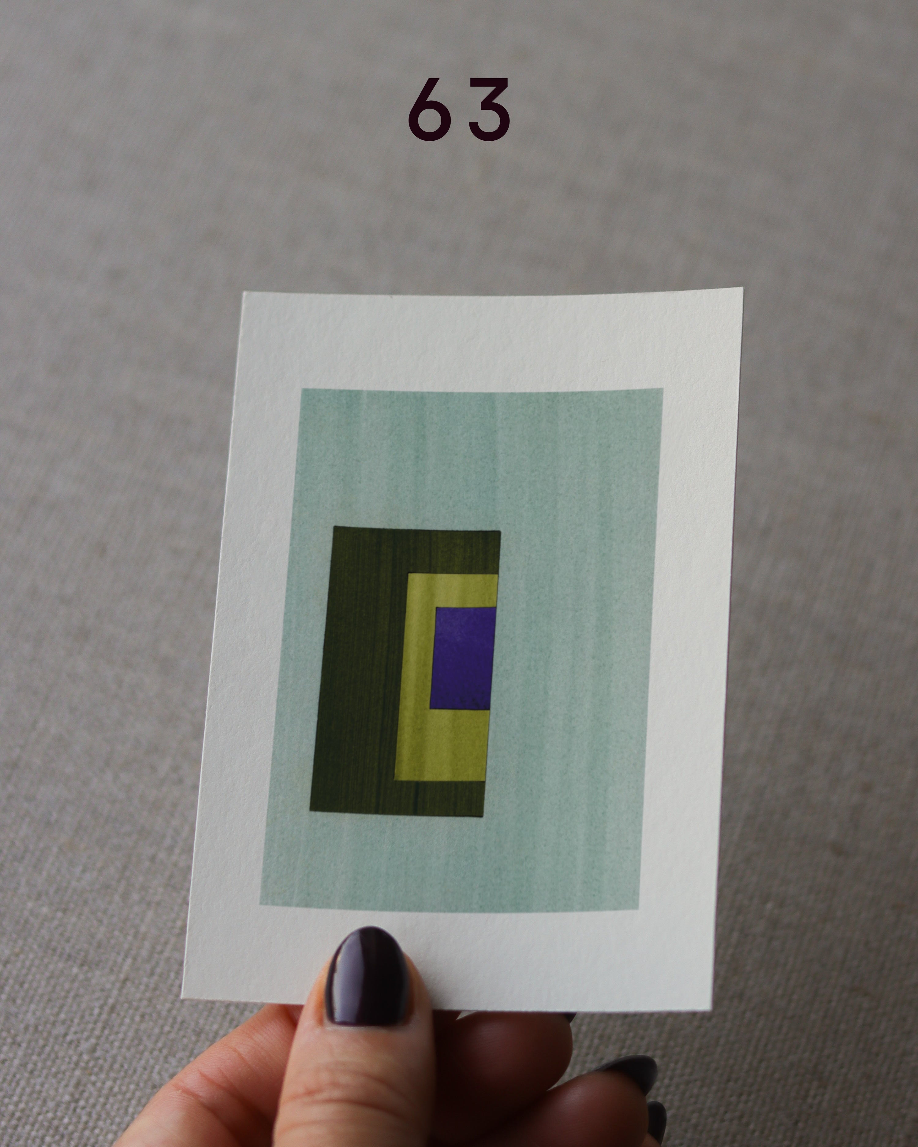 Hand holding sample of a turquoise, olive green, yellow and blue minimalist abstract art print with a geometric design over a grey linen background