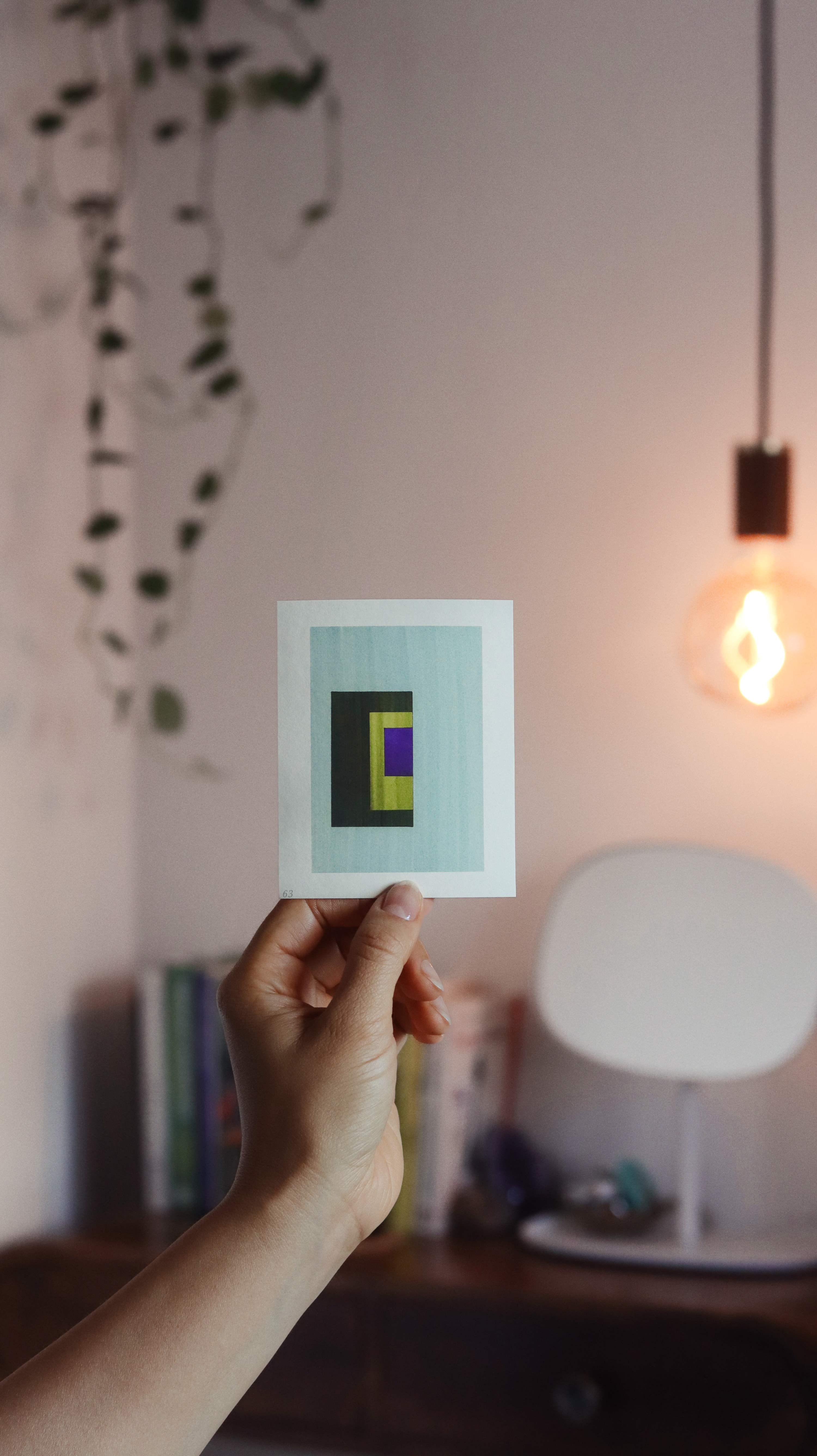 Hand holding sample of a turquoise and blue minimalist abstract art print with a geometric design in a peaceful, modern bedroom with plants and pendant light