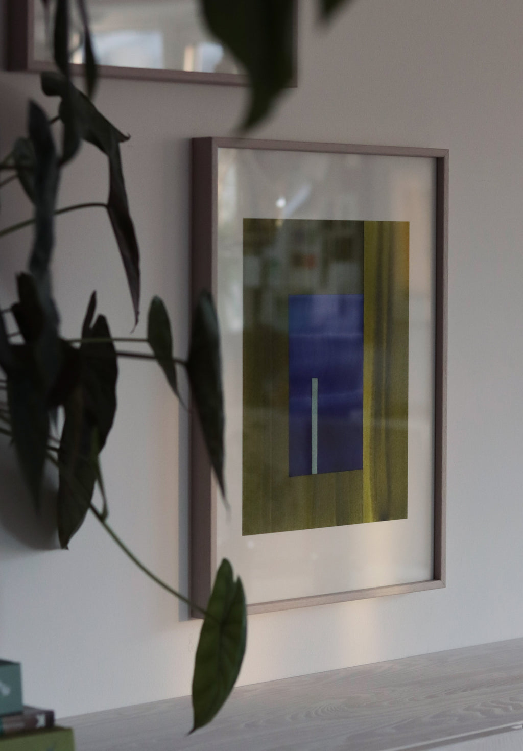 Framed green and blue geometric abstract artwork on a wall with plant leaves in the foreground