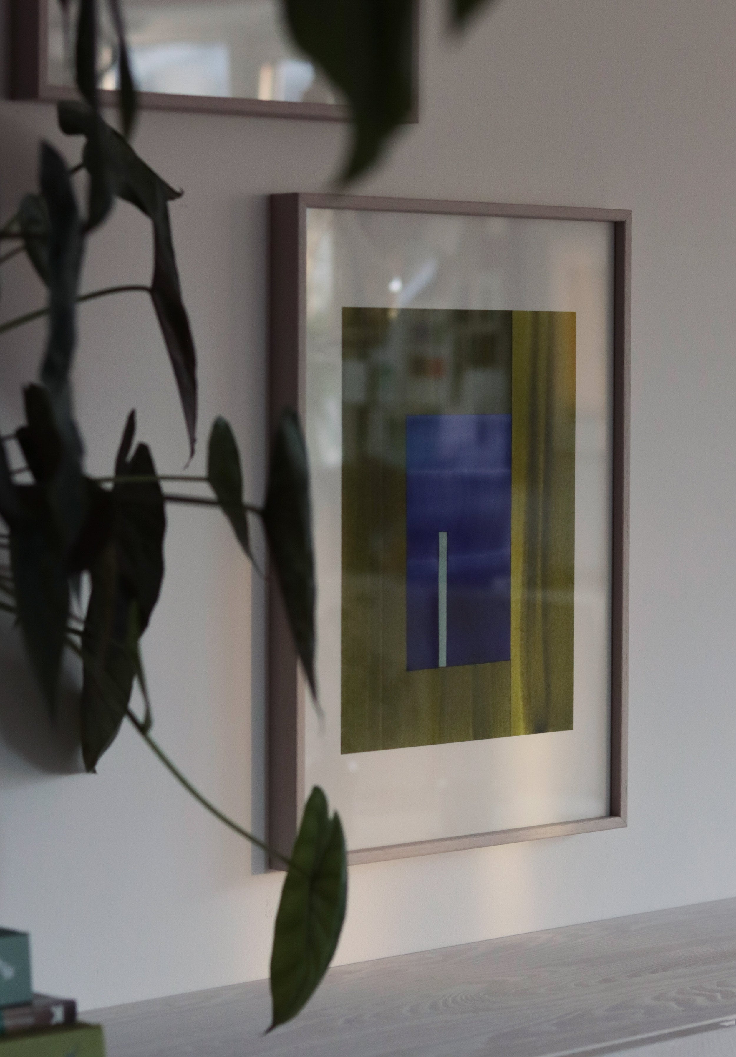 Framed green and blue geometric abstract artwork on a wall with plant leaves in the foreground