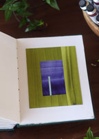 Open sketchbook with a green and blue geometric abstract art piece on a dark wooden table with tubes of watercolour paint and plants.