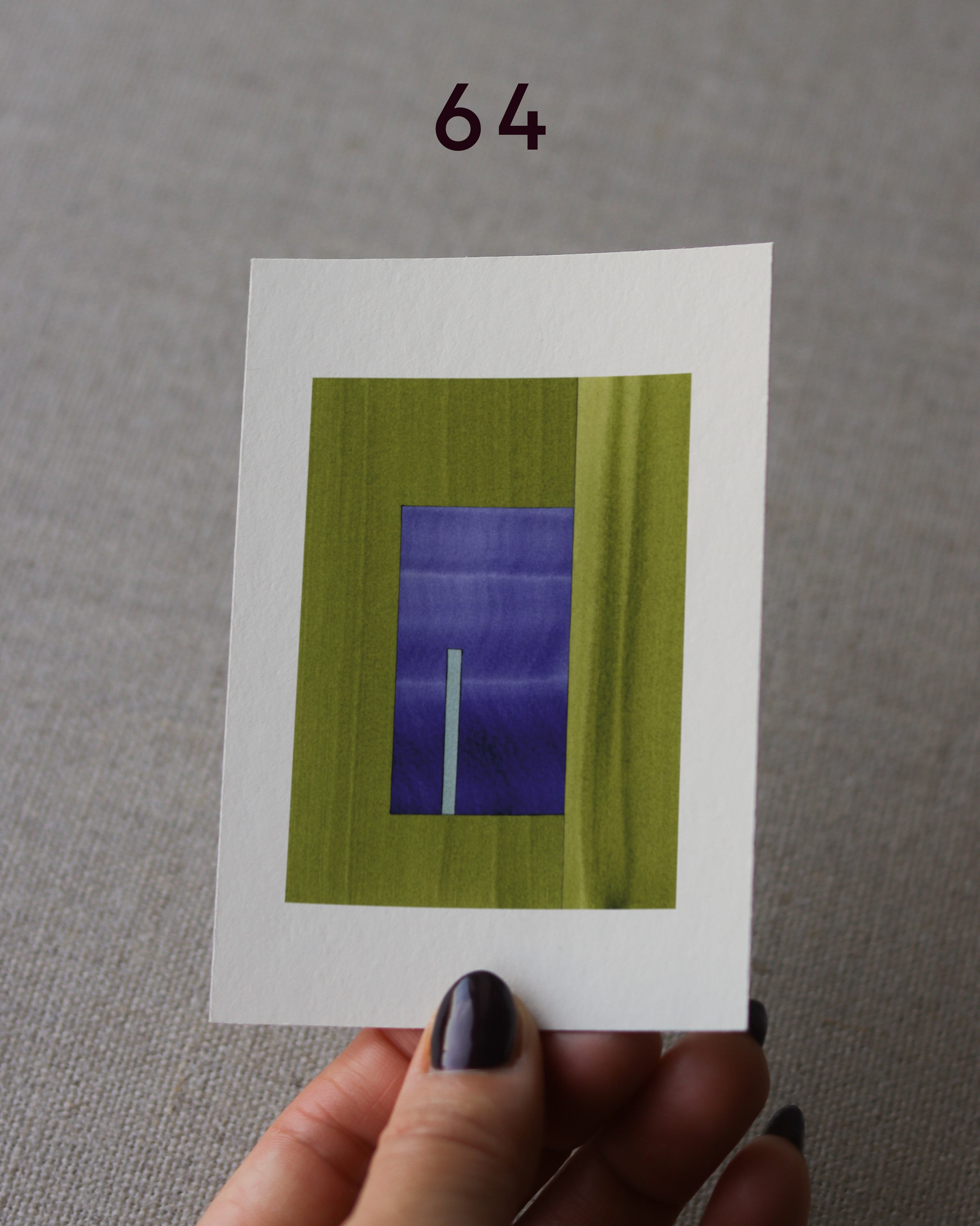 Hand holding sample of a green and blue minimalist abstract art print with a geometric design over a grey linen background