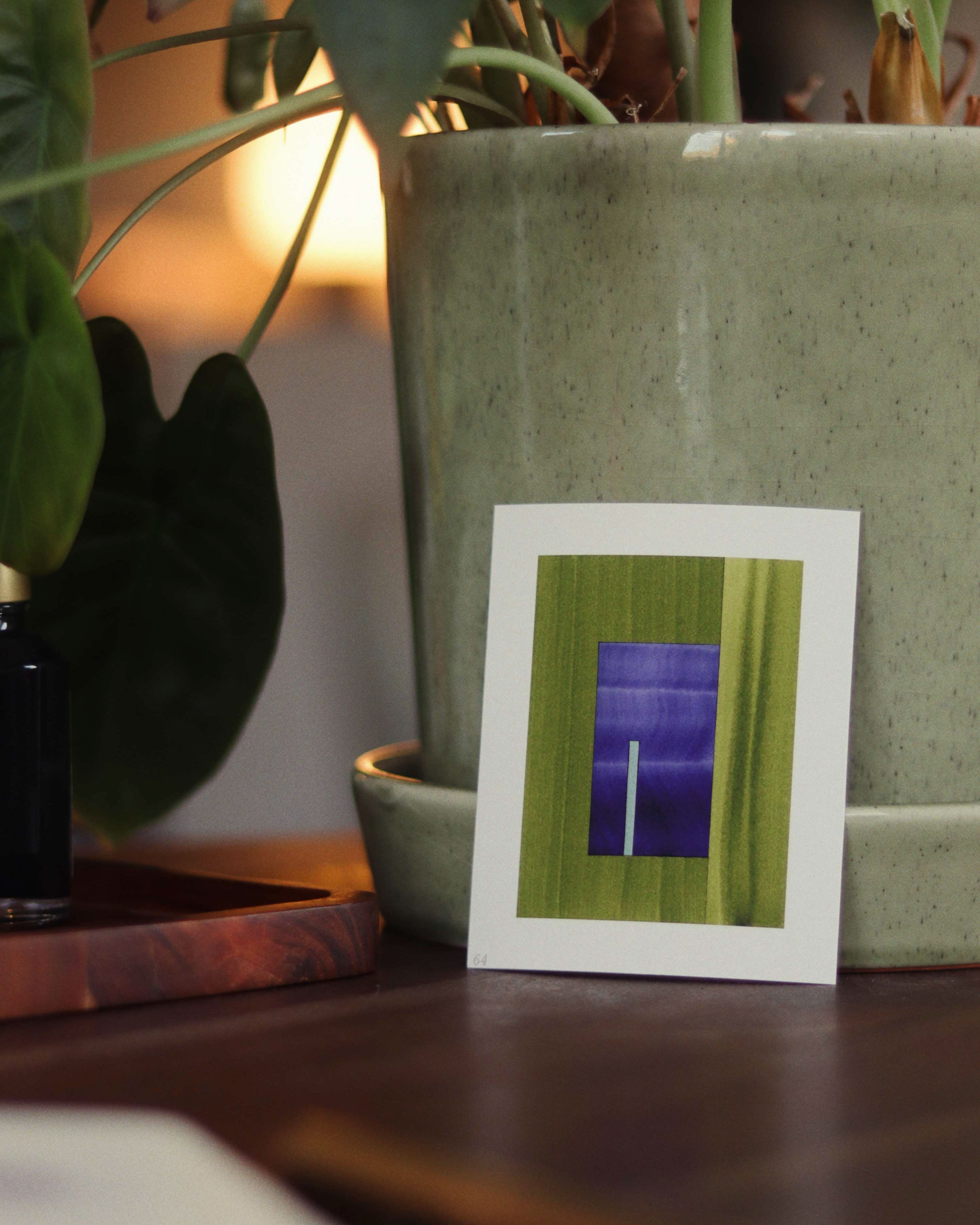 Small blue and green geometric abstract art print sample sitting on a dark wooden desk in front of a potted plant