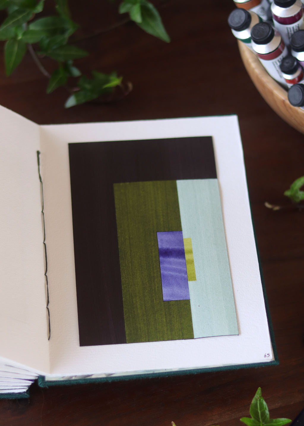 Open sketchbook with a geometric abstract art piece on a dark wooden table with tubes of watercolour paint and plants.