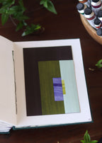 Open sketchbook with a geometric abstract art piece on a dark wooden table with tubes of watercolour paint and plants.