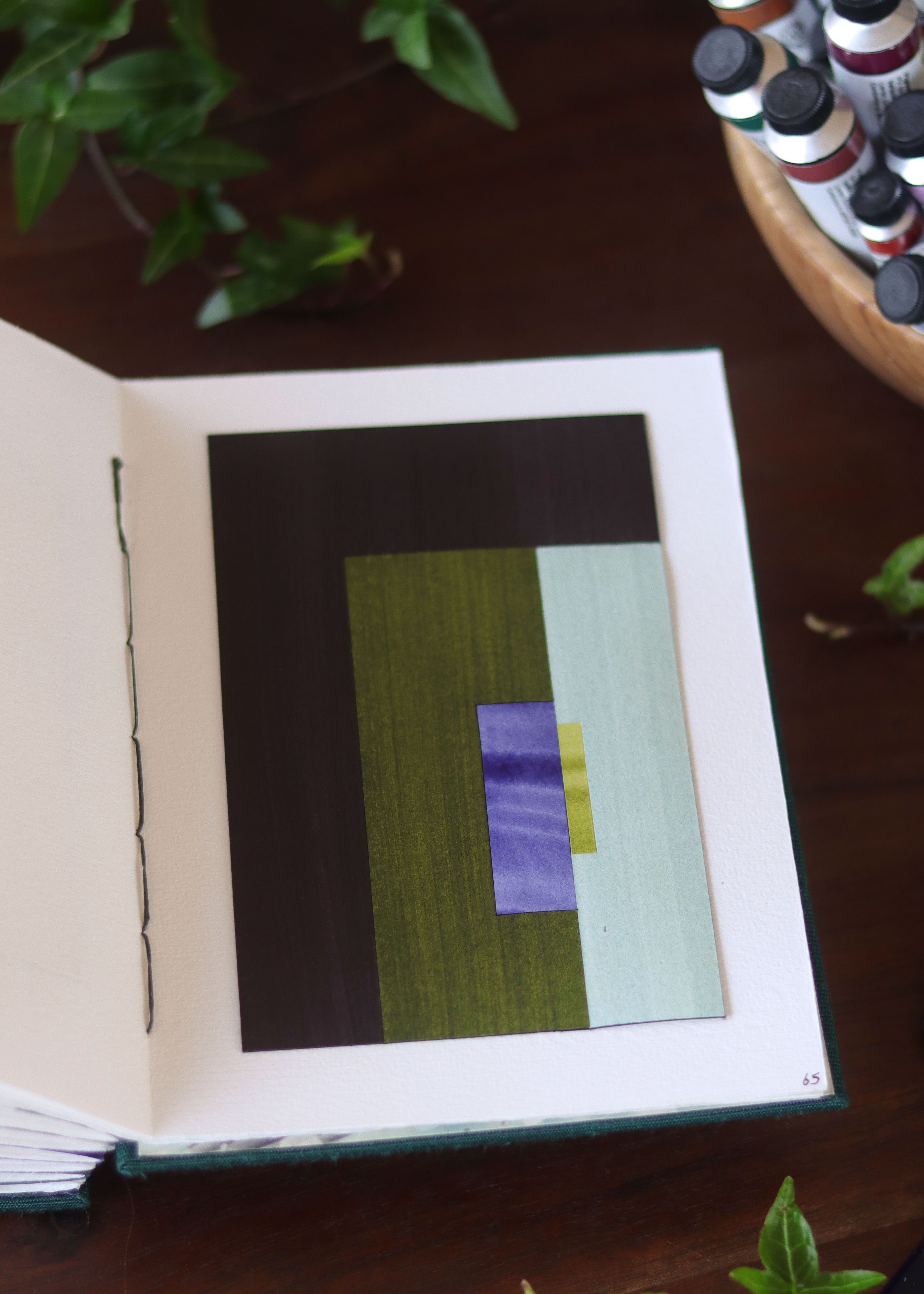 Open sketchbook with a geometric abstract art piece on a dark wooden table with tubes of watercolour paint and plants.